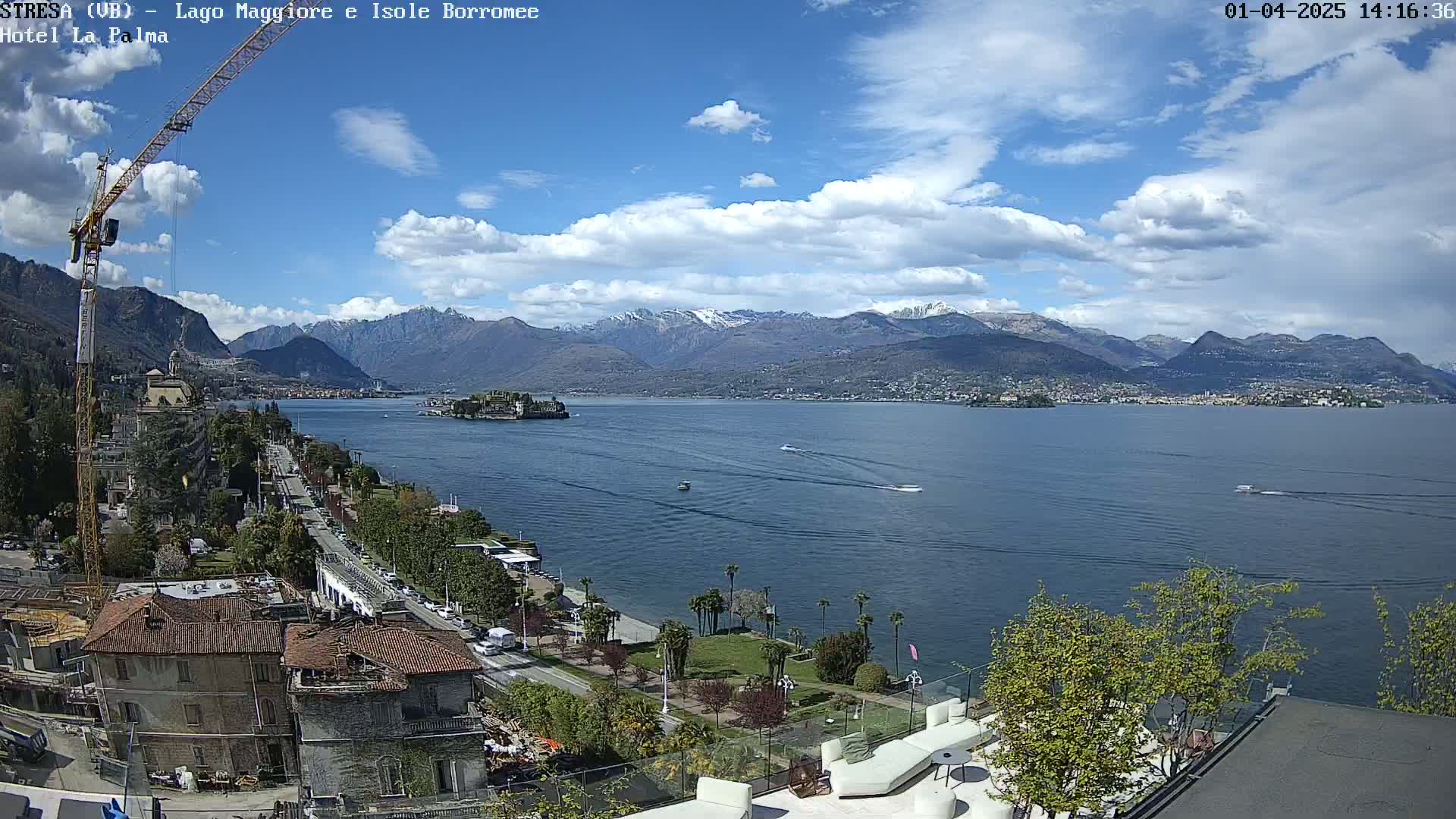 A partly cloudy day shows a large lake with boats, a shoreline with buildings and trees, a construction crane, and snow-capped mountains in the distance.