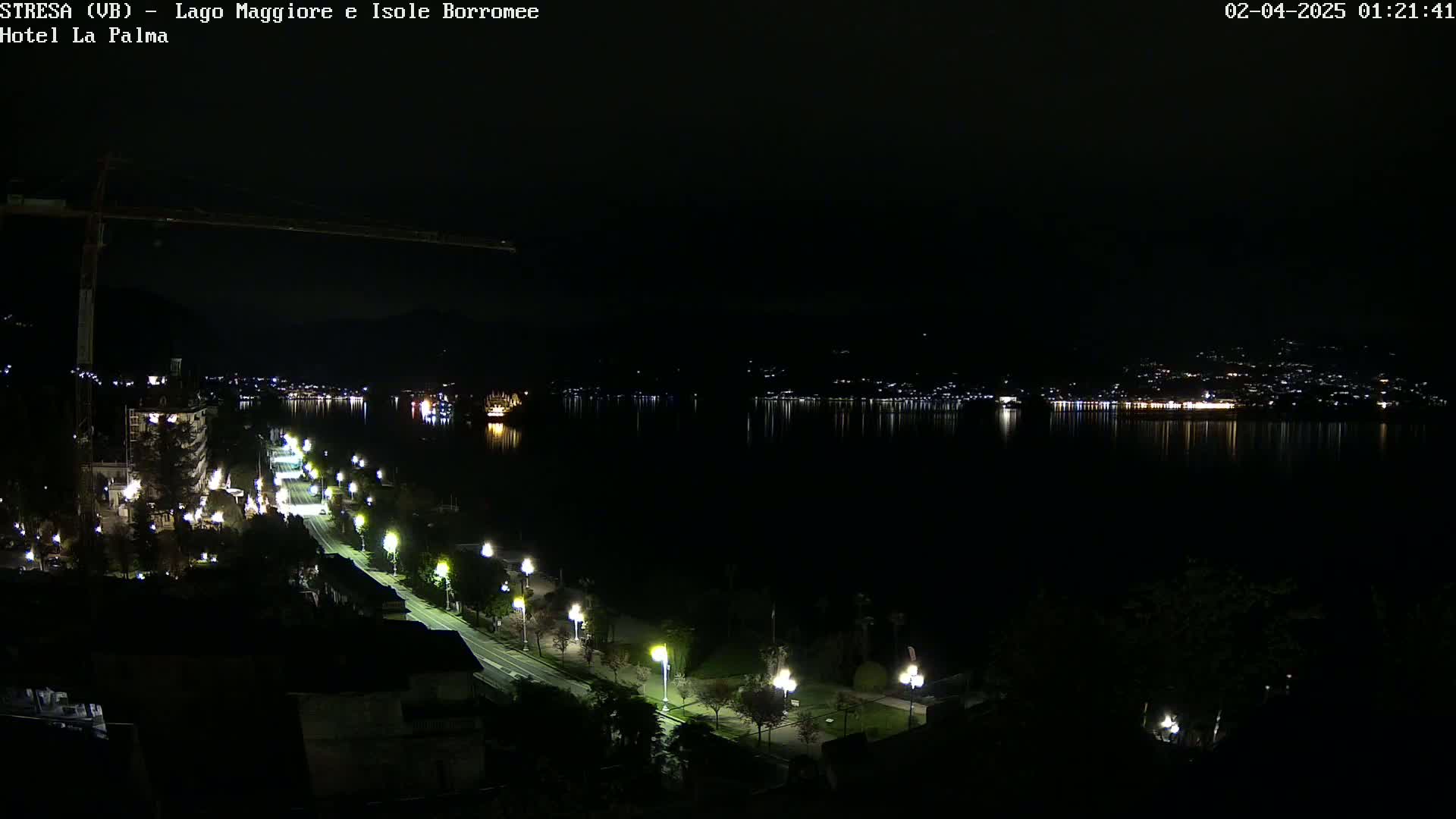 A nighttime view of a city and lake, with a road lined with lights leading to the water's edge, where the lights of buildings across the lake are reflected in the calm water.