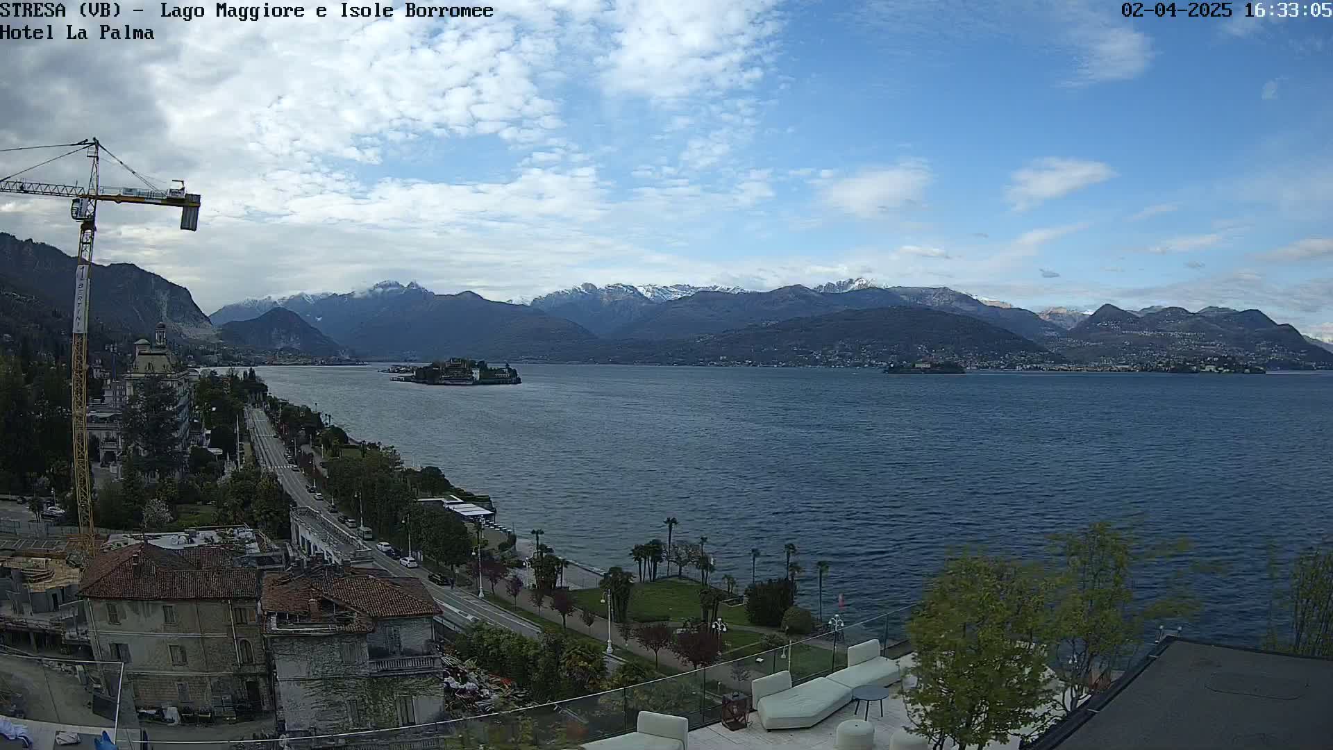 A partly cloudy day reveals a panoramic view of a lake with snow-capped mountains in the distance, a small island in the middle, and a lakeside town with buildings and a crane visible.