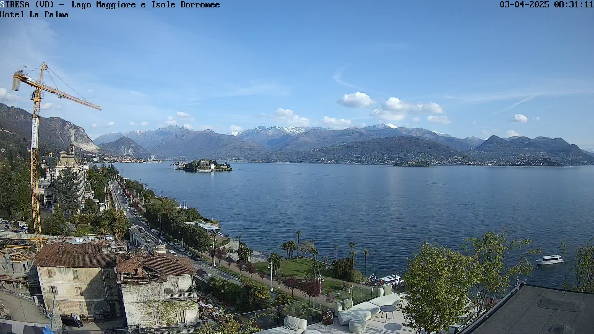 A partly sunny day overlooks a lake with a small island in the middle, mountains in the background, and a cityscape along the shoreline with a construction crane visible.