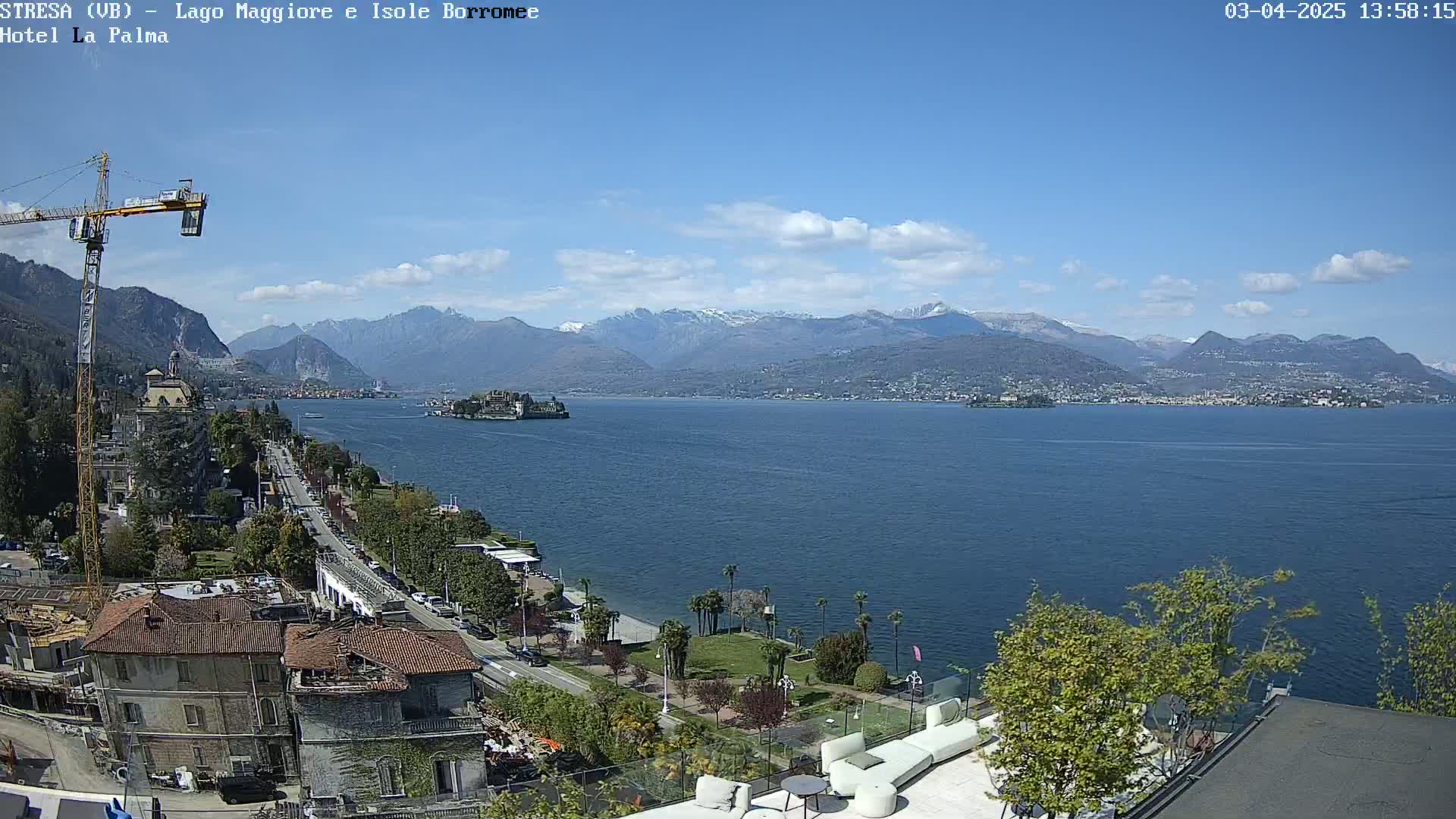 A partly sunny day overlooking a large lake with snow-capped mountains in the distance, a town along the shore, and a construction crane visible in the foreground.