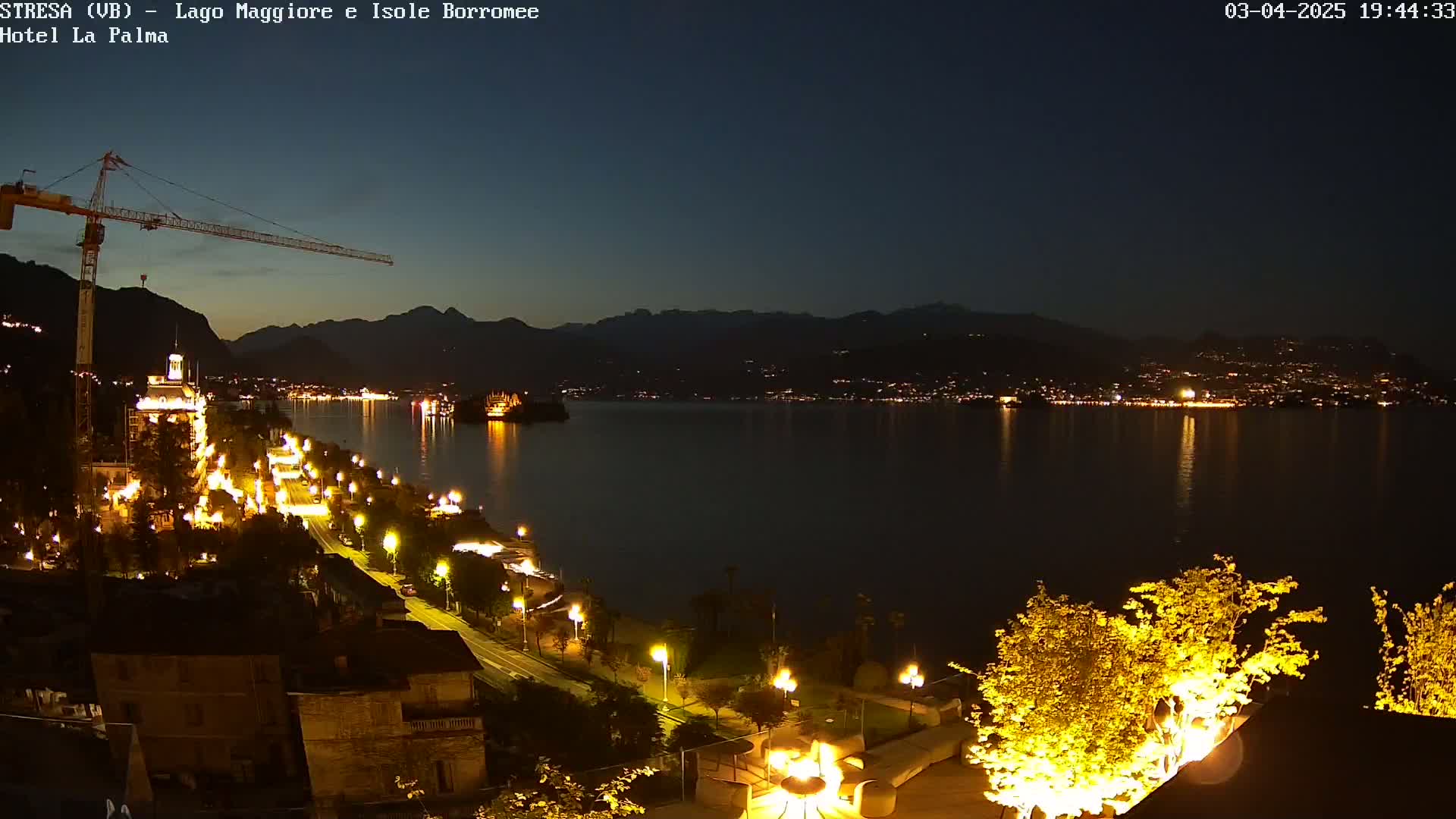 A nighttime view of a lakeside town illuminated with lights, featuring a construction crane, a calm lake reflecting the lights, and mountains in the background under a dark, clear sky.