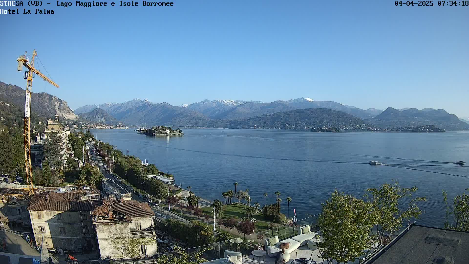A clear, sunny day overlooks a lake with snow-capped mountains in the background, a small island in the middle of the lake, and a cityscape along the shoreline with a construction crane.