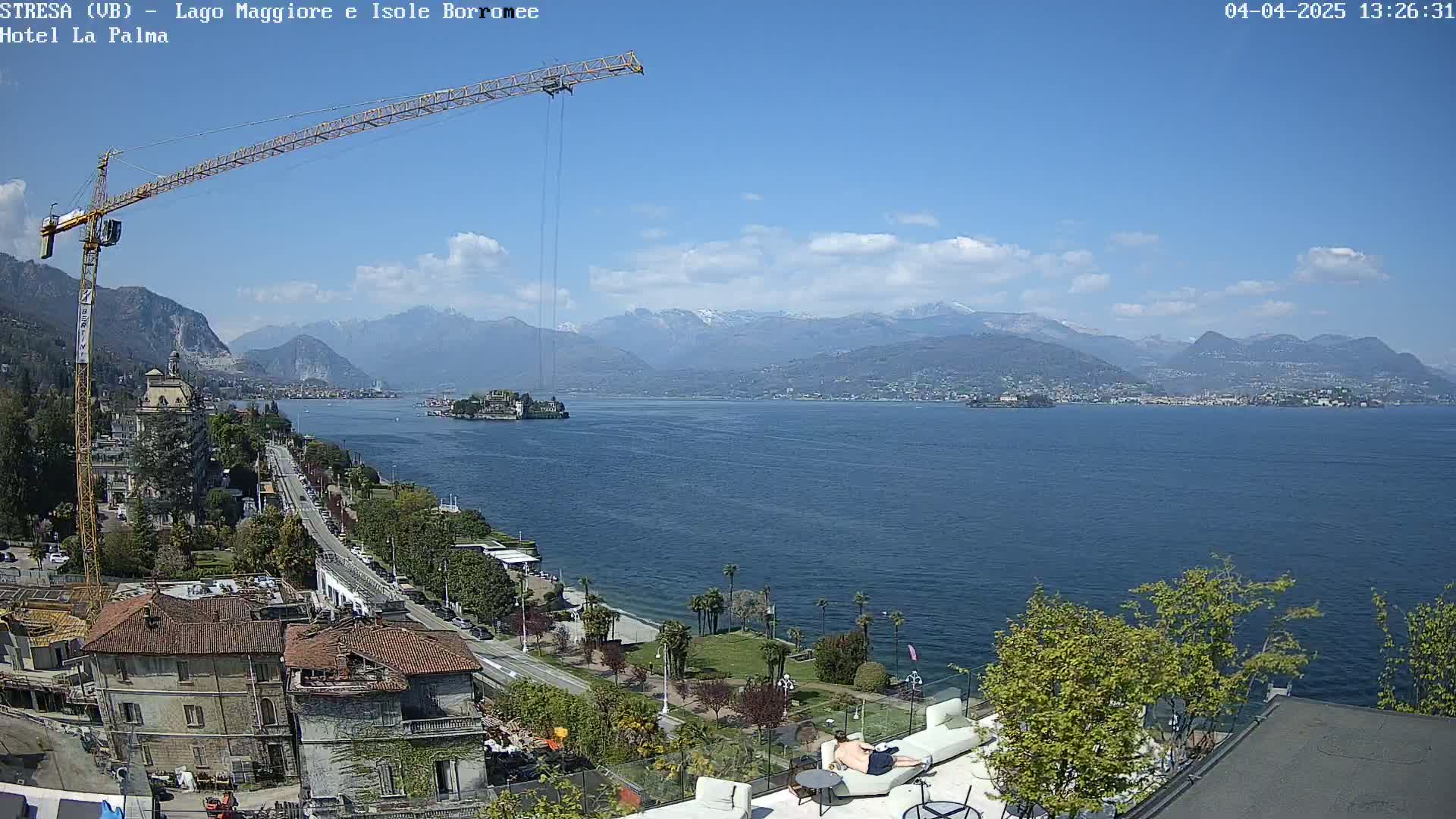 A large construction crane overlooks a lake with mountains in the background under a mostly sunny sky.