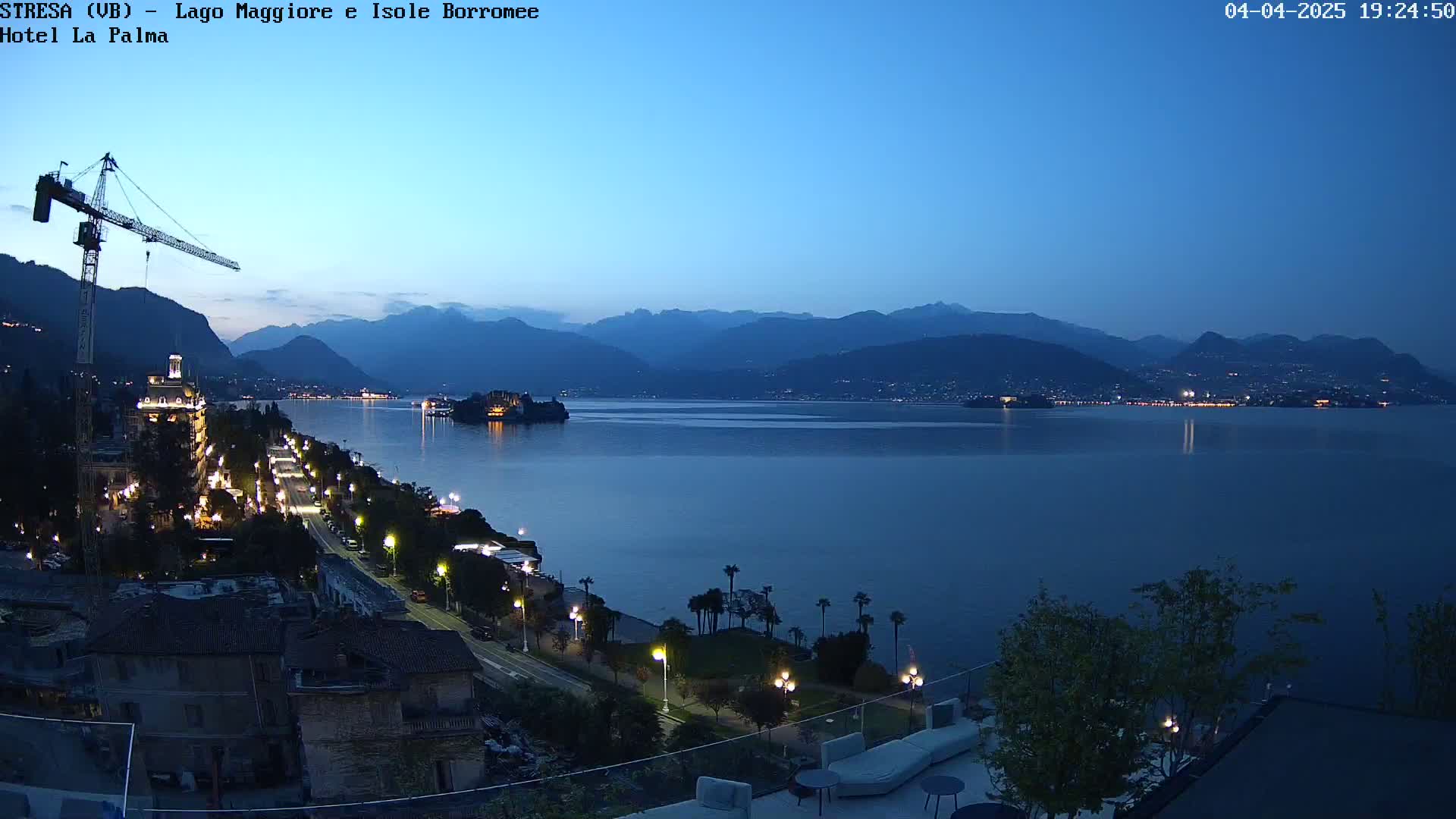 A twilight view of a lake, mountains, and a lakeside town with a construction crane, illuminated by streetlights.