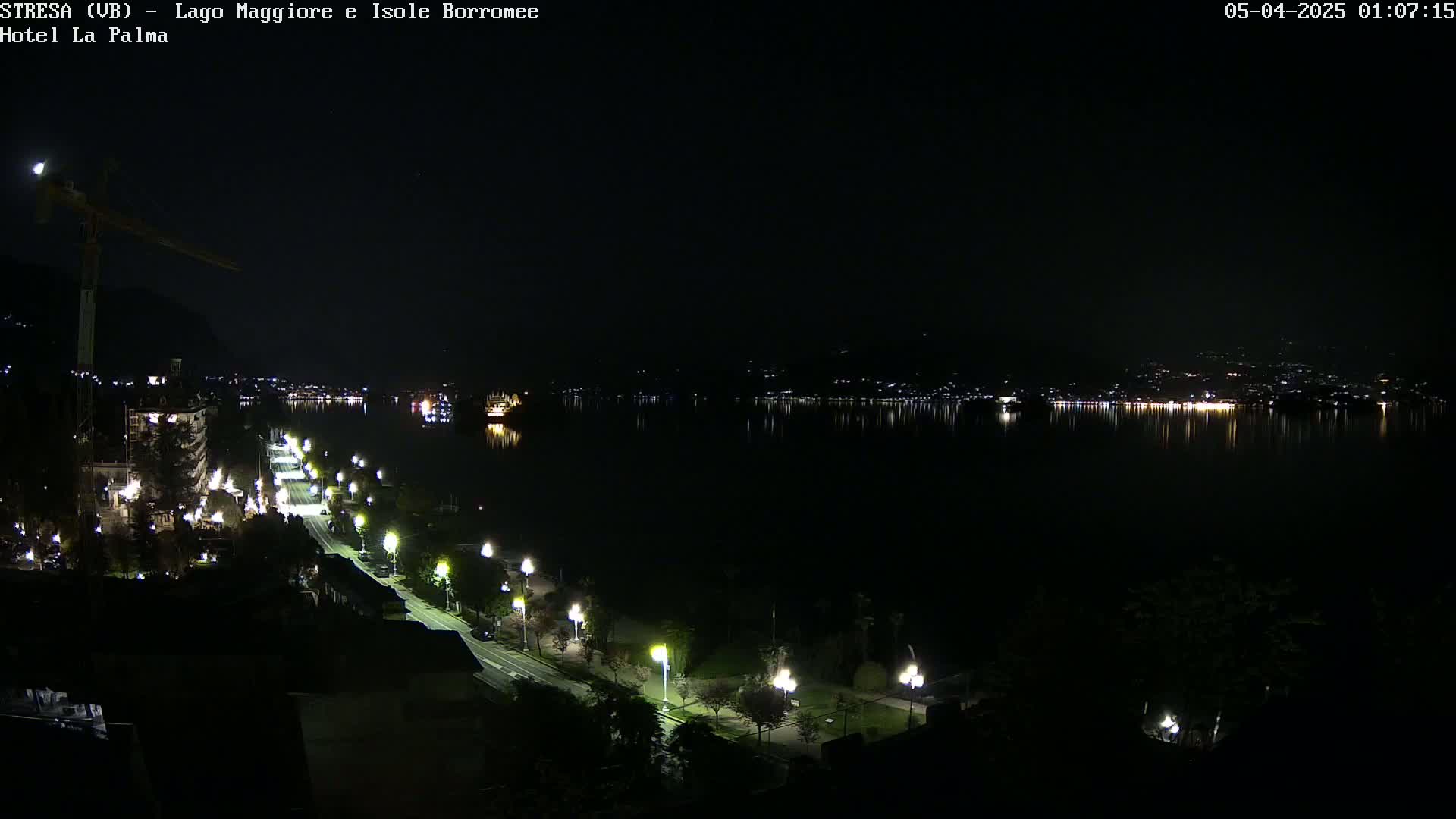 A nighttime view of a city's waterfront, featuring a calm lake reflecting the lights of buildings across the water, with a road lined with streetlights leading down to the shore.