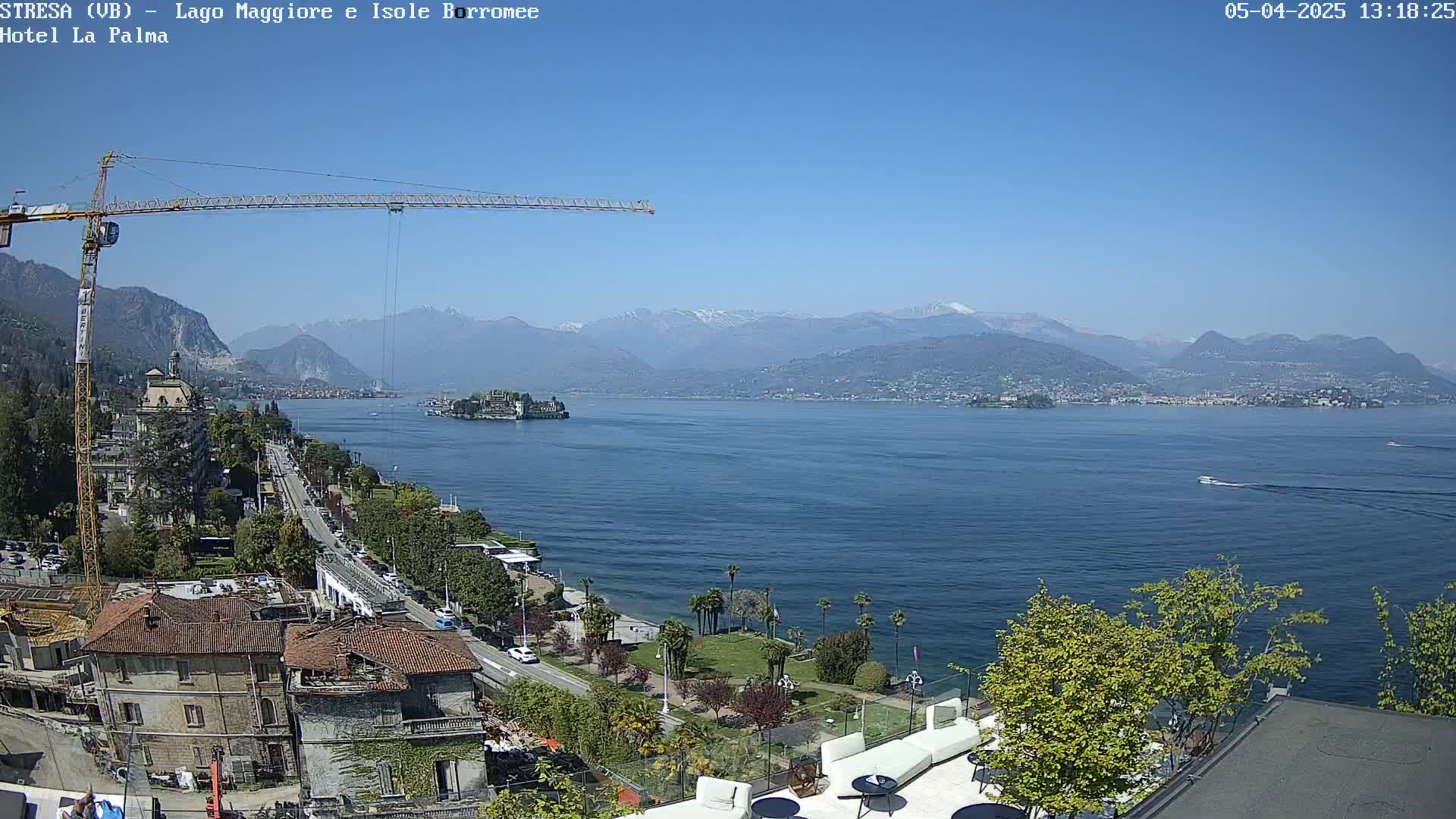 A large body of water with a small island and distant mountains under a clear blue sky is viewed from an elevated position, showing buildings, trees, and a construction crane in the foreground.