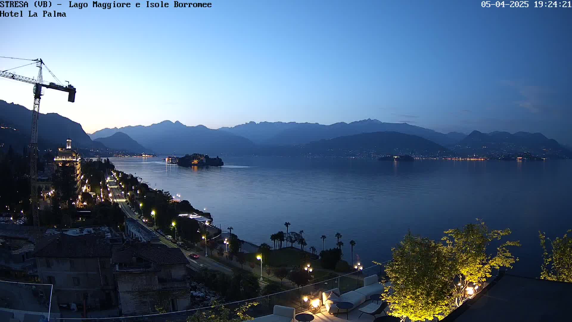 A twilight view shows a calm lake with mountains in the background, a town with lights along the shoreline, and a construction crane in the foreground.
