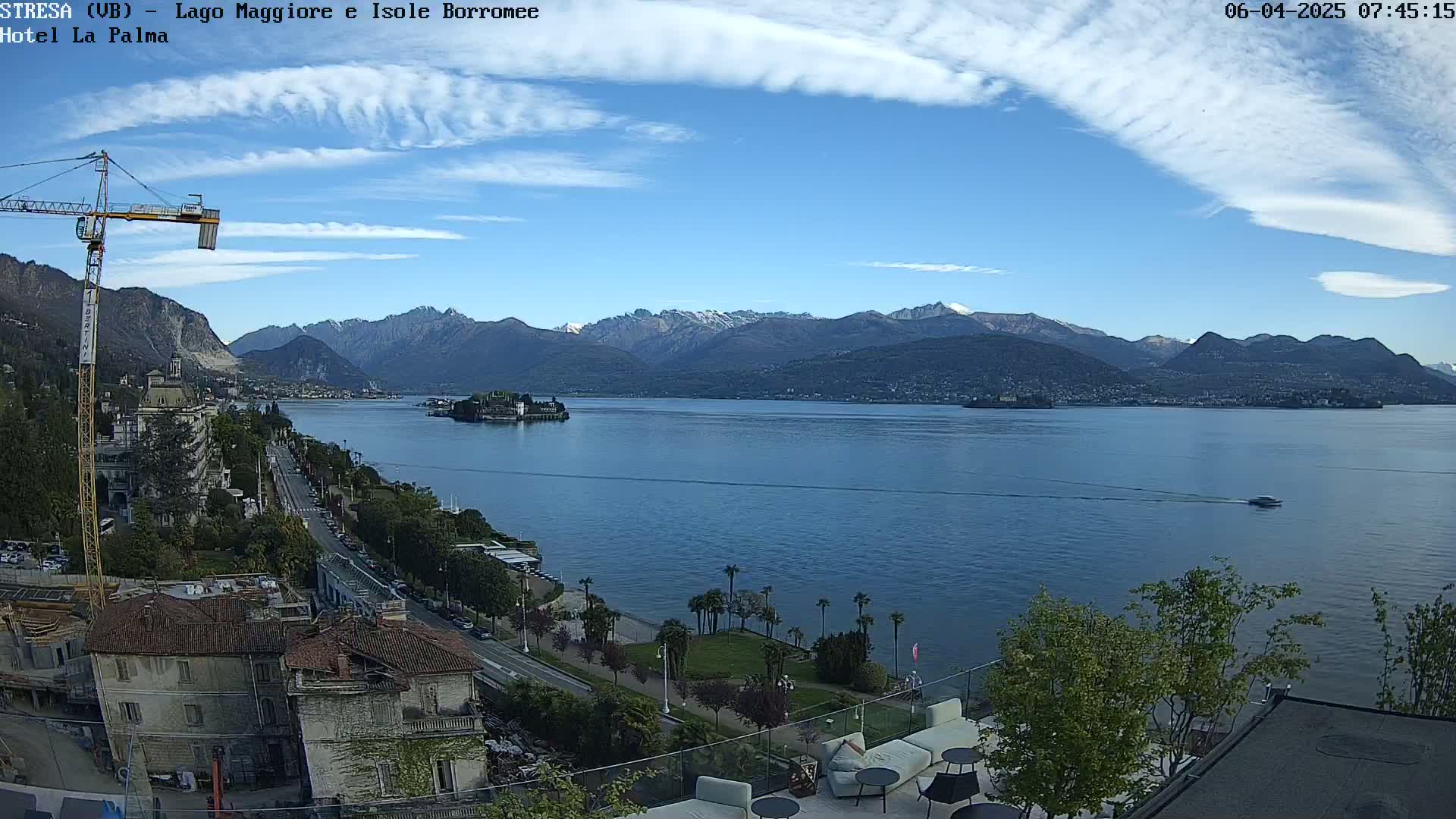 A partly cloudy sky overlooks a calm lake with mountains in the background, a small island, buildings along the shore, and a boat on the water.