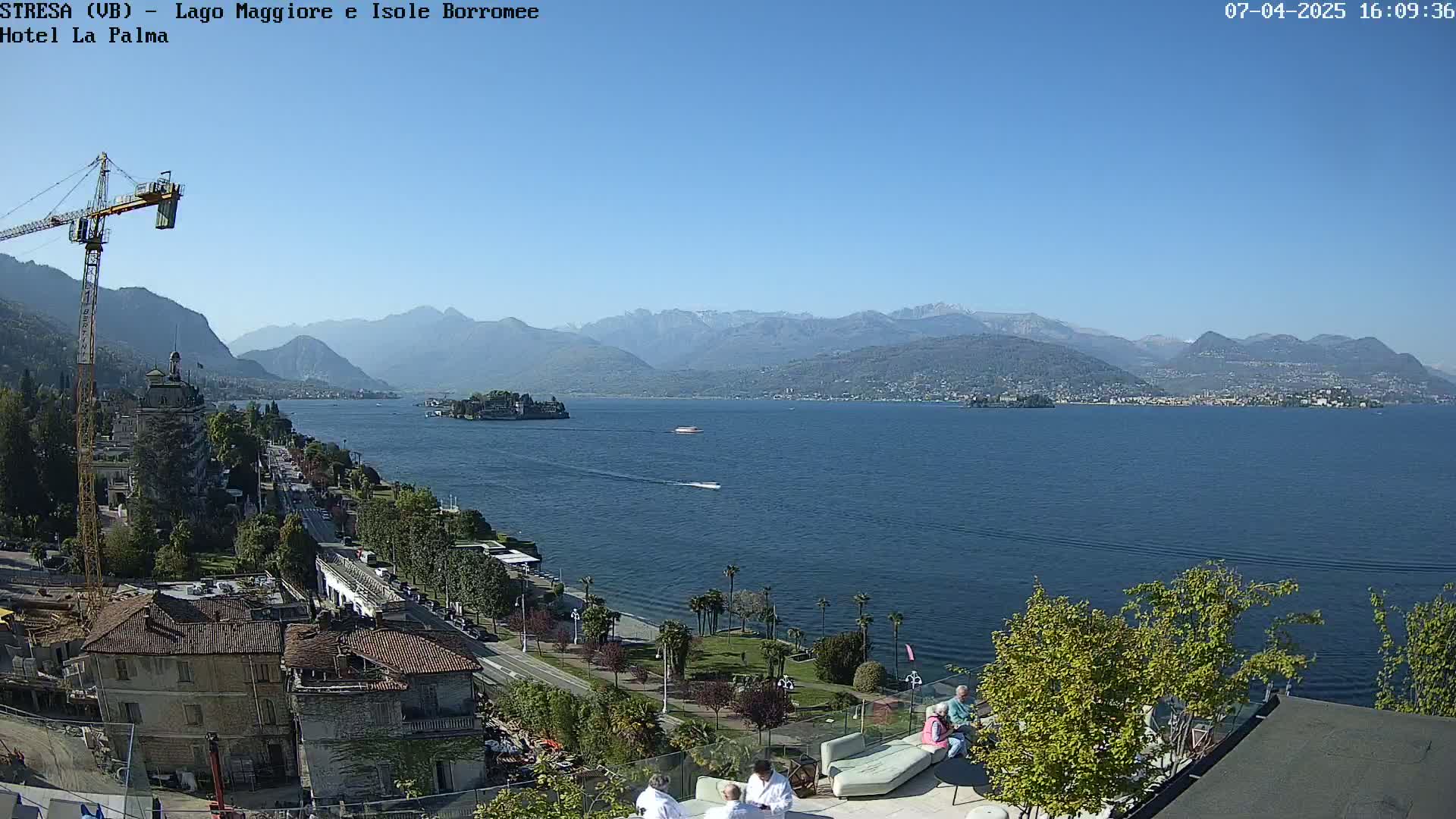 A sunny view of a large lake, with mountains in the background, a small island in the middle of the lake, buildings along the shore, and several people sitting on a patio.