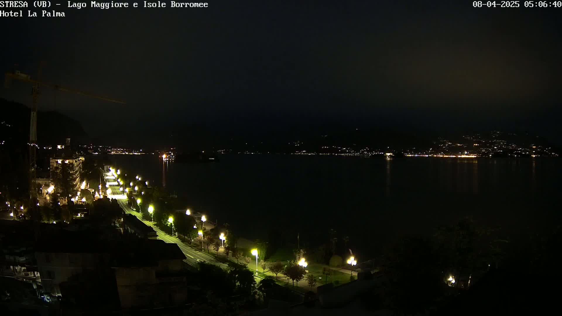 A nighttime view of a city illuminated by streetlights lining a road beside a dark body of water, with a construction crane visible on the left and a distant town across the water.
