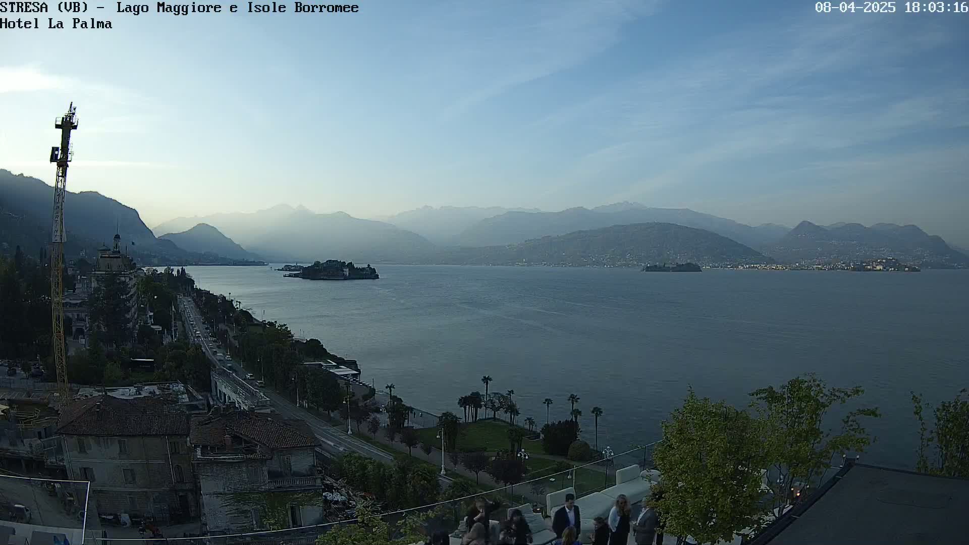 A panoramic view of a lake with mountains in the background under a clear, twilight sky, shows a town along the shore with people gathered near a building overlooking the water.