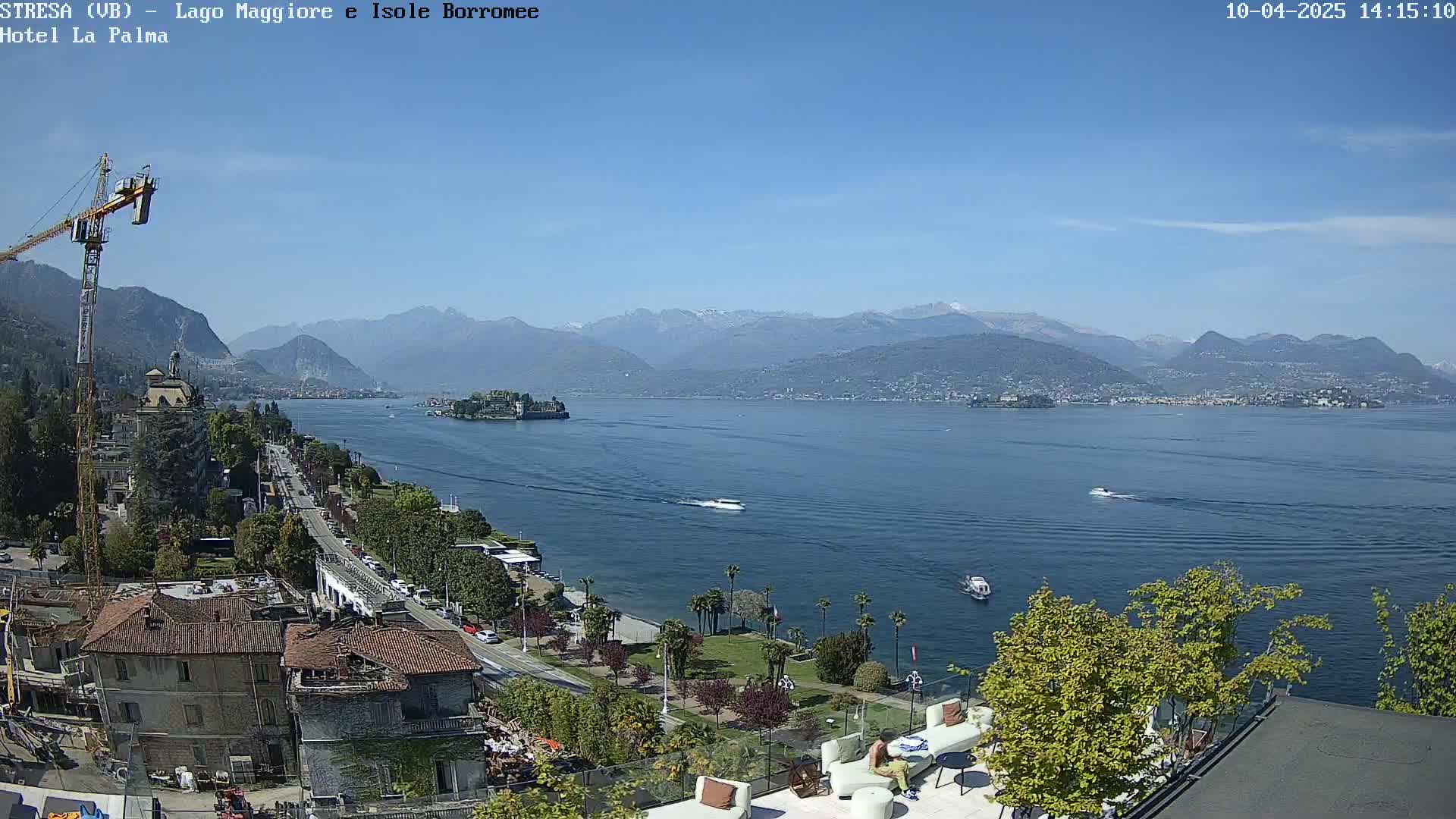 A sunny day overlooks a calm lake with mountains in the distance, several boats on the water, buildings and trees along the shoreline, and a construction crane near the foreground.