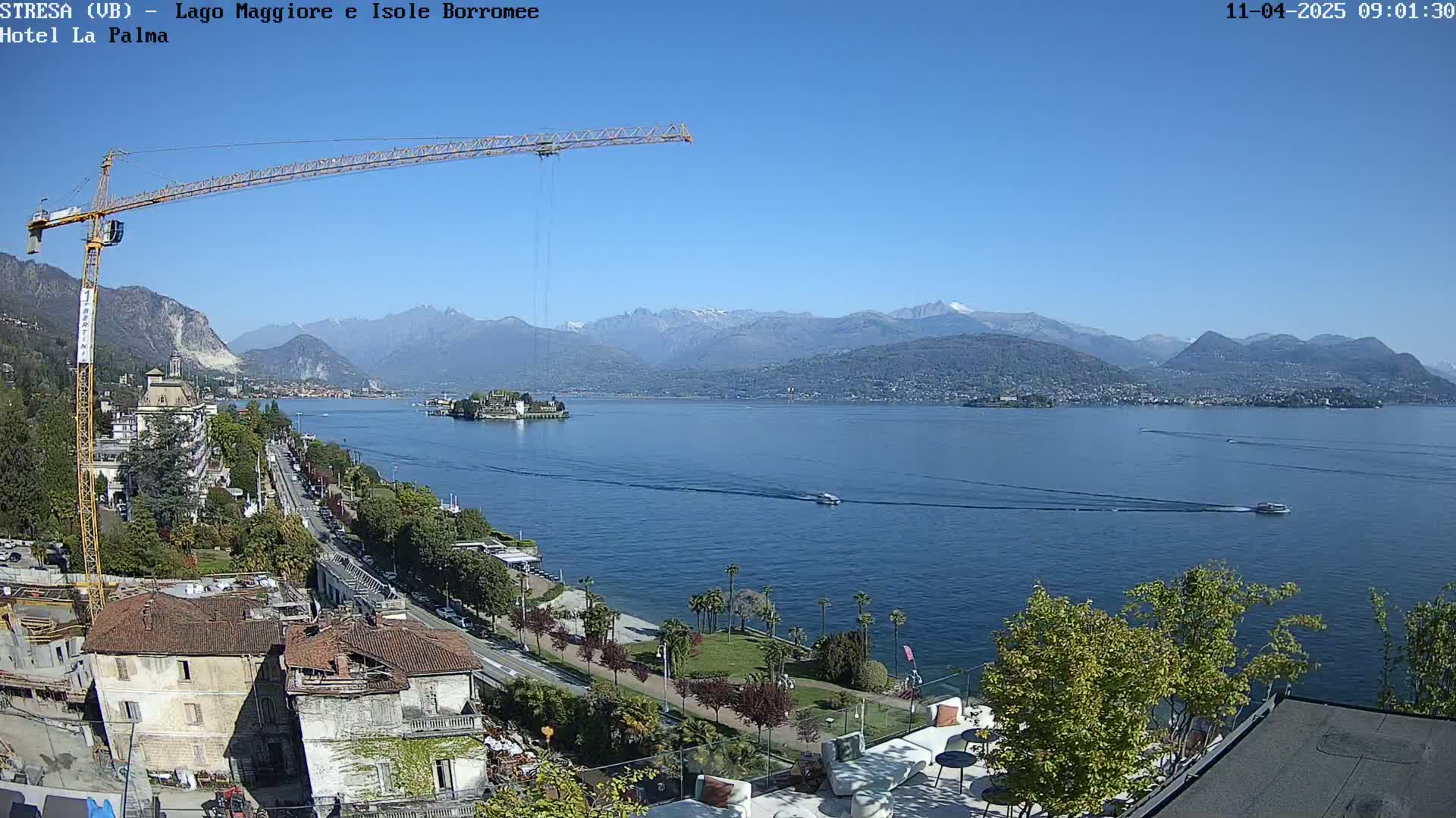 A large lake with mountains in the background is seen from an elevated vantage point on a clear sunny day, with a crane visible in the foreground near buildings and a waterfront road.