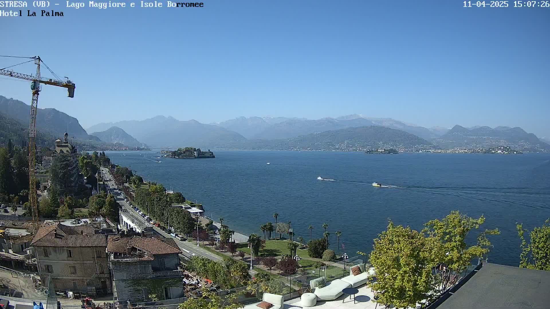 A sunny view of a large lake with mountains in the background, a small island in the middle of the lake, and a town with buildings and a road along the shore.