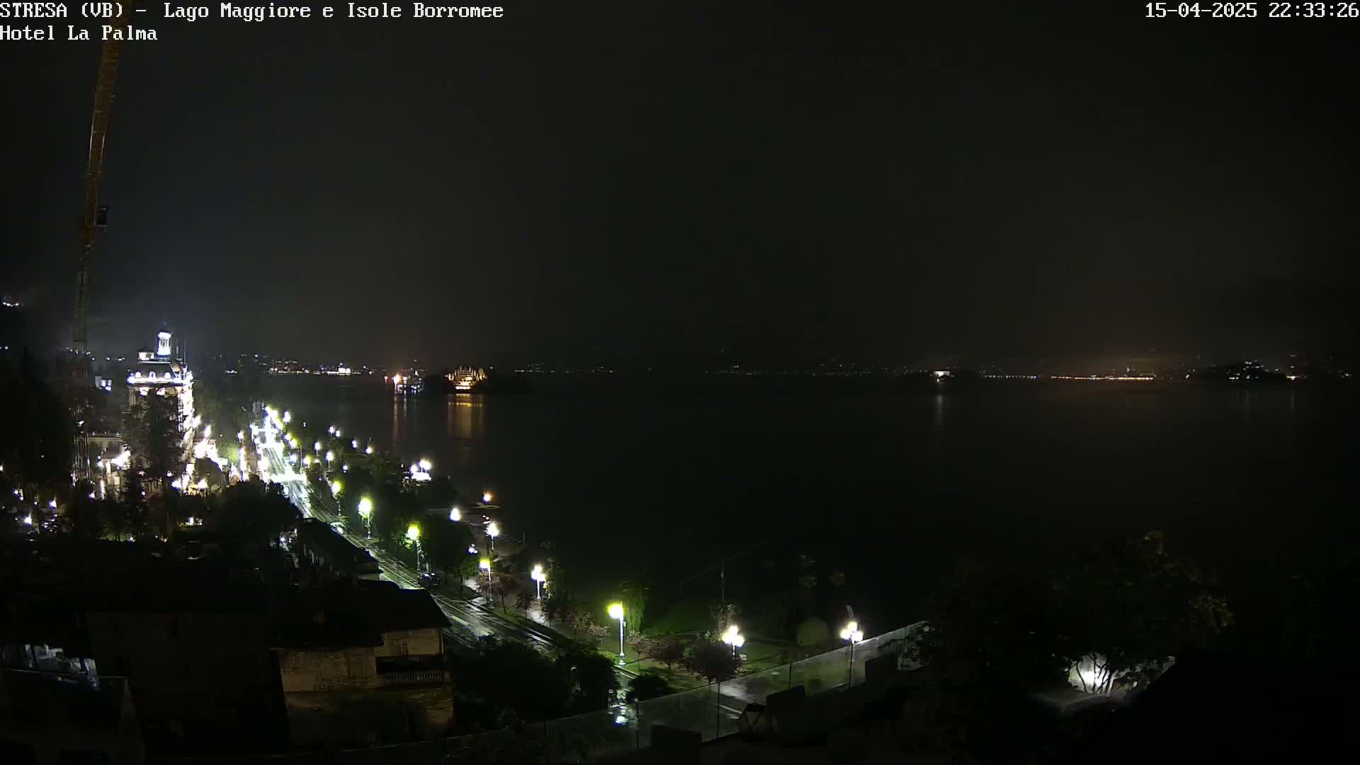 A nighttime view of a rain-slicked lakeside road with illuminated buildings and streetlights, overlooking a dark lake with faintly visible lights on the opposite shore.