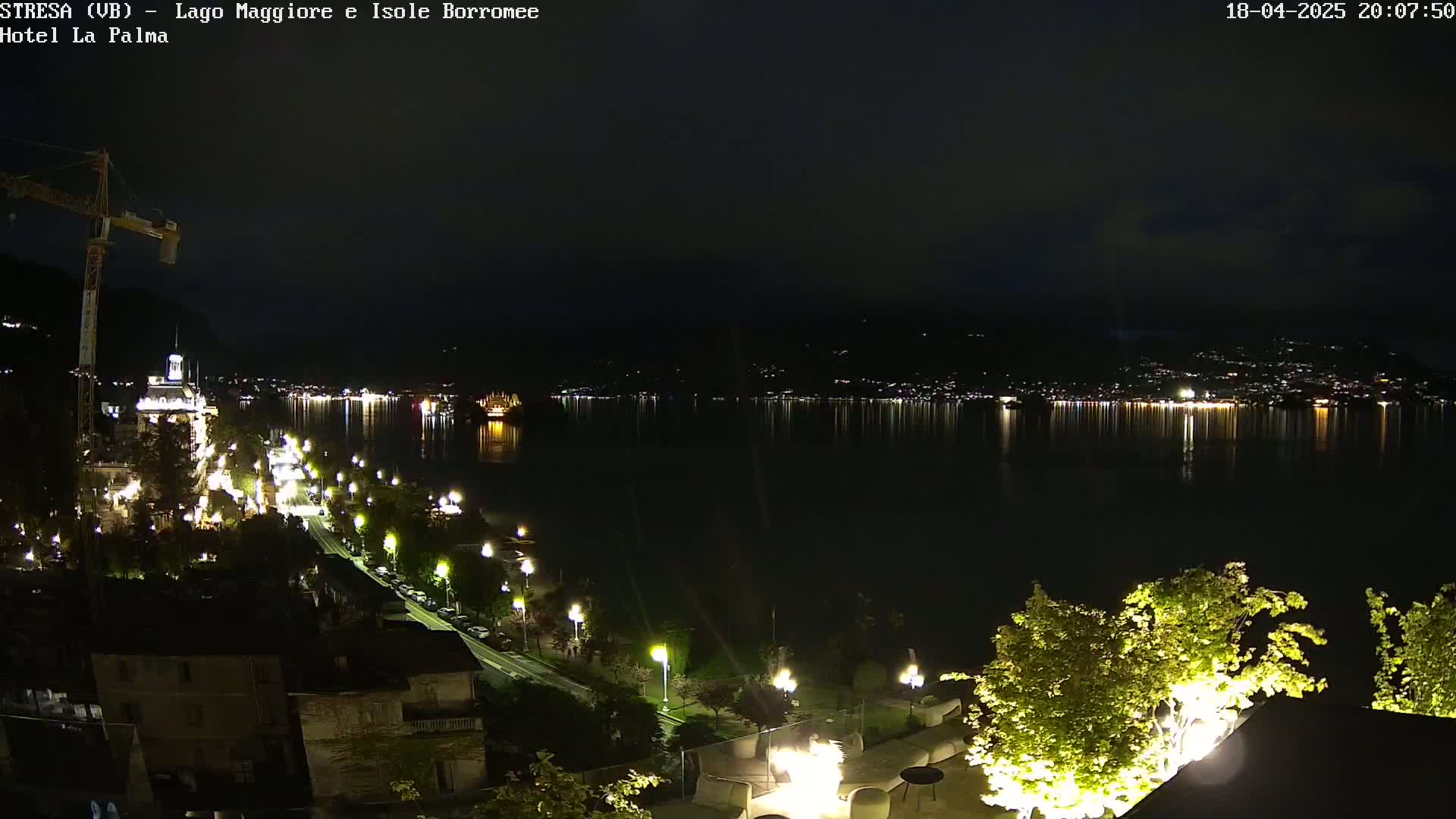 A nighttime view of a lakefront town with illuminated buildings and streets, extending to a calm, dark lake reflecting the lights of the opposite shore under a mostly clear sky.