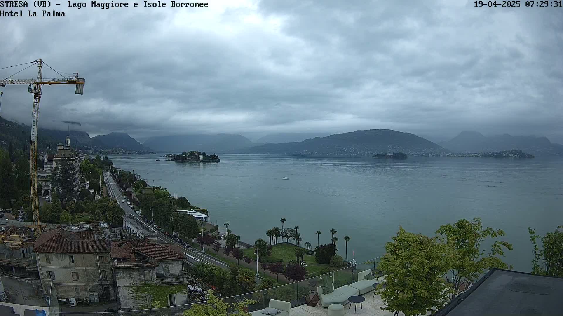 A cloudy day overlooks a calm lake with distant mountains and a small island, viewed from an elevated position showing buildings and a road along the lakeshore.