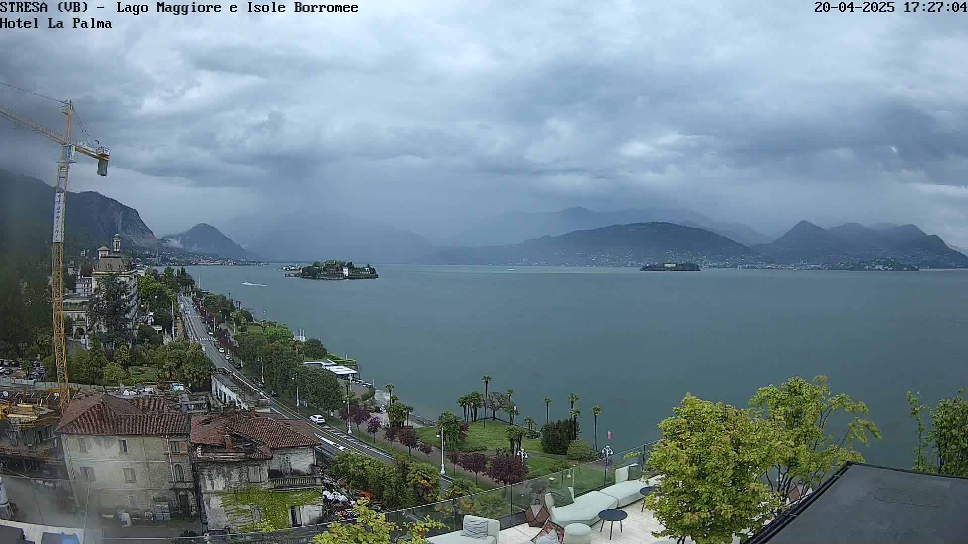 A large lake is seen under a cloudy sky, with mountains in the background and a town along the shoreline, including a construction crane and a small island in the lake.