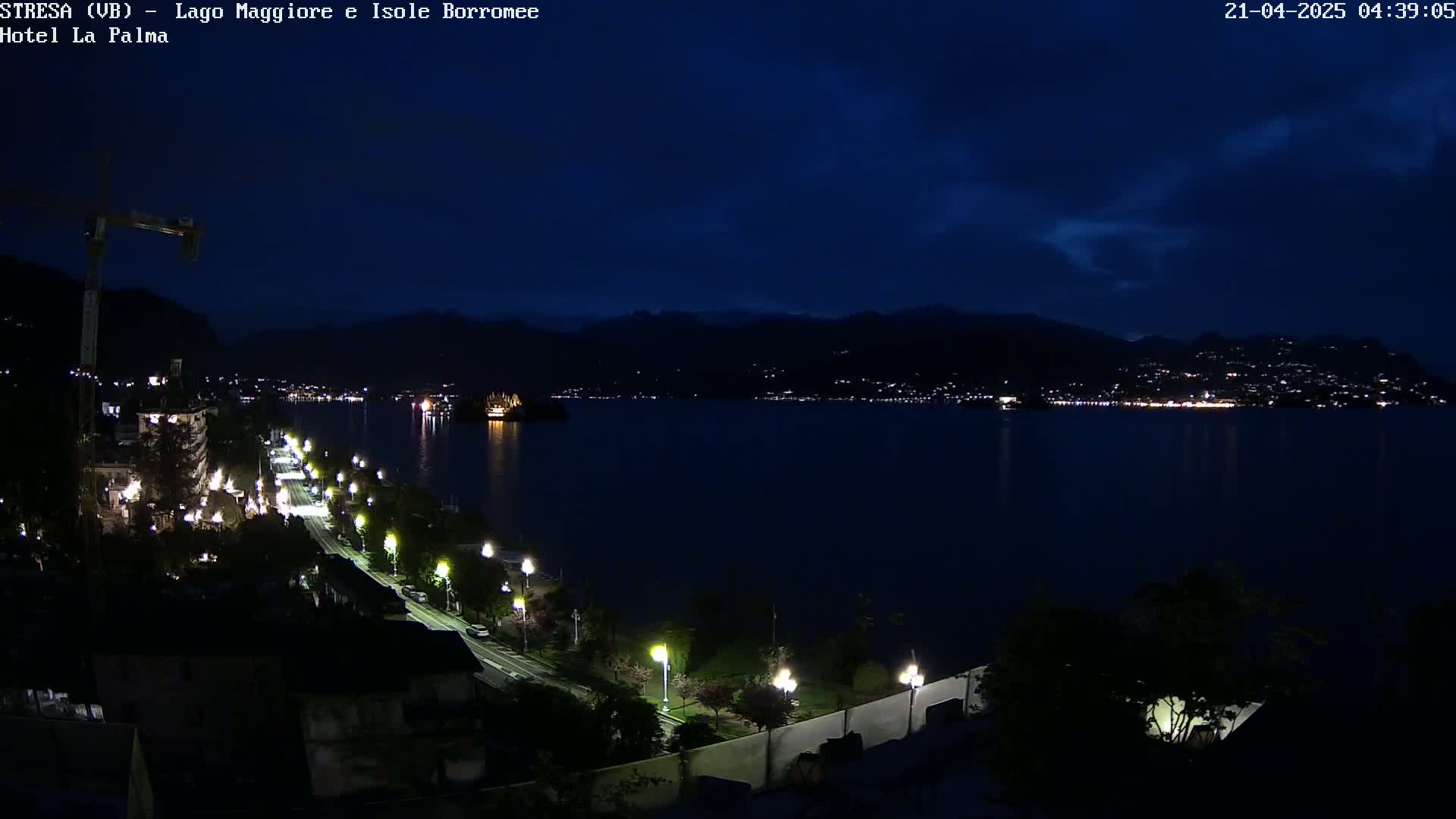 A nighttime view of a lake with a city and mountains in the background, under a dark, cloudy sky, showing a road lit by streetlights leading down to the water.