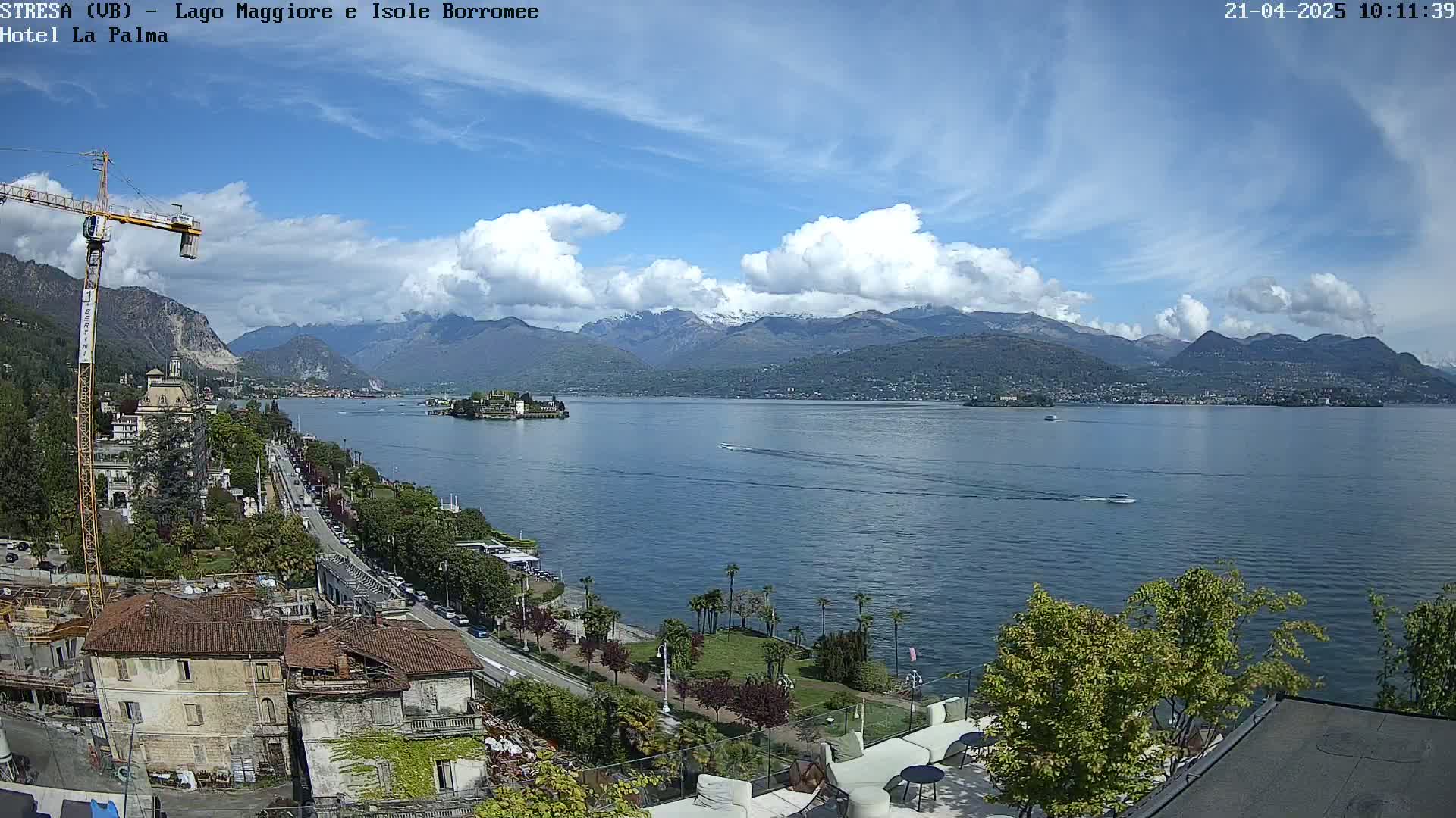 A partly cloudy day shows a view of a lake with mountains in the background, a small island in the middle, and buildings and trees along the shoreline.