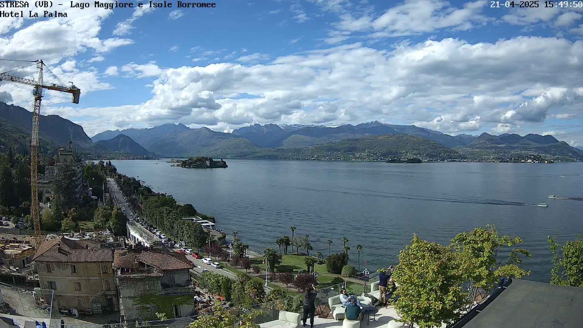 A partly cloudy day overlooking a large lake with mountains in the background, a town visible along the lakeshore, and several people sitting on a patio.