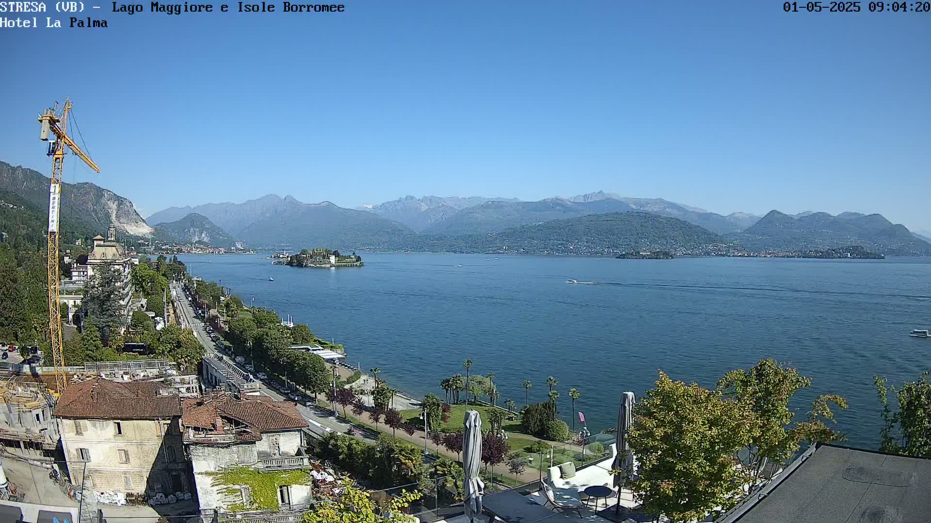 A sunny view overlooking a calm lake, bordered by buildings, trees, and mountains in the distance, with a construction crane visible near the foreground.