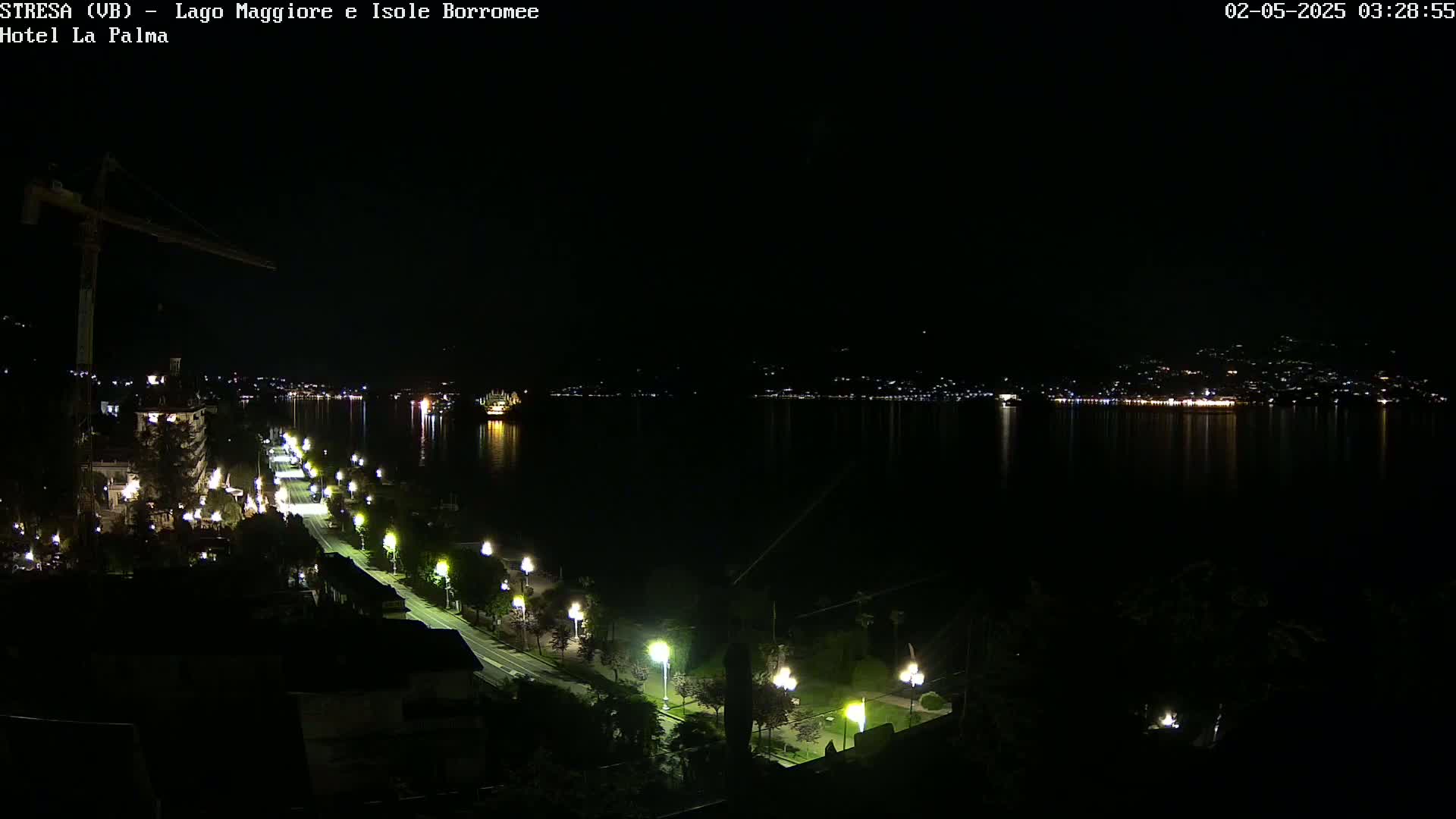 A nighttime view of a city along a body of water, with a road lined with lights leading toward a distant, illuminated shoreline.