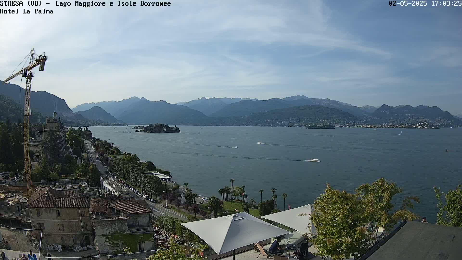 A sunny day overlooks a calm lake with mountains in the background, buildings and trees lining the shore, and a few boats on the water.