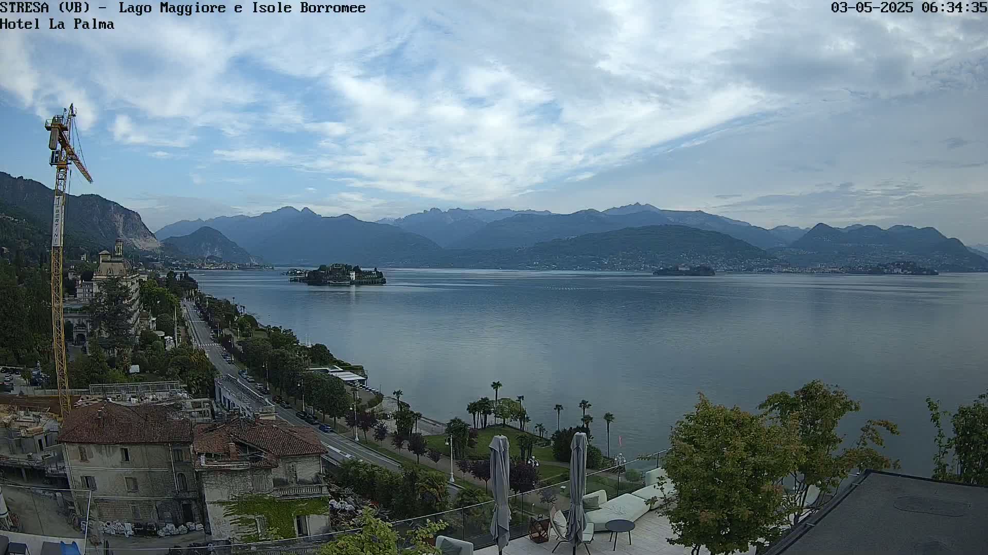 A partly cloudy day reveals a tranquil lake scene, with mountains in the distance and a cityscape along the shore, featuring a construction crane and palm trees.