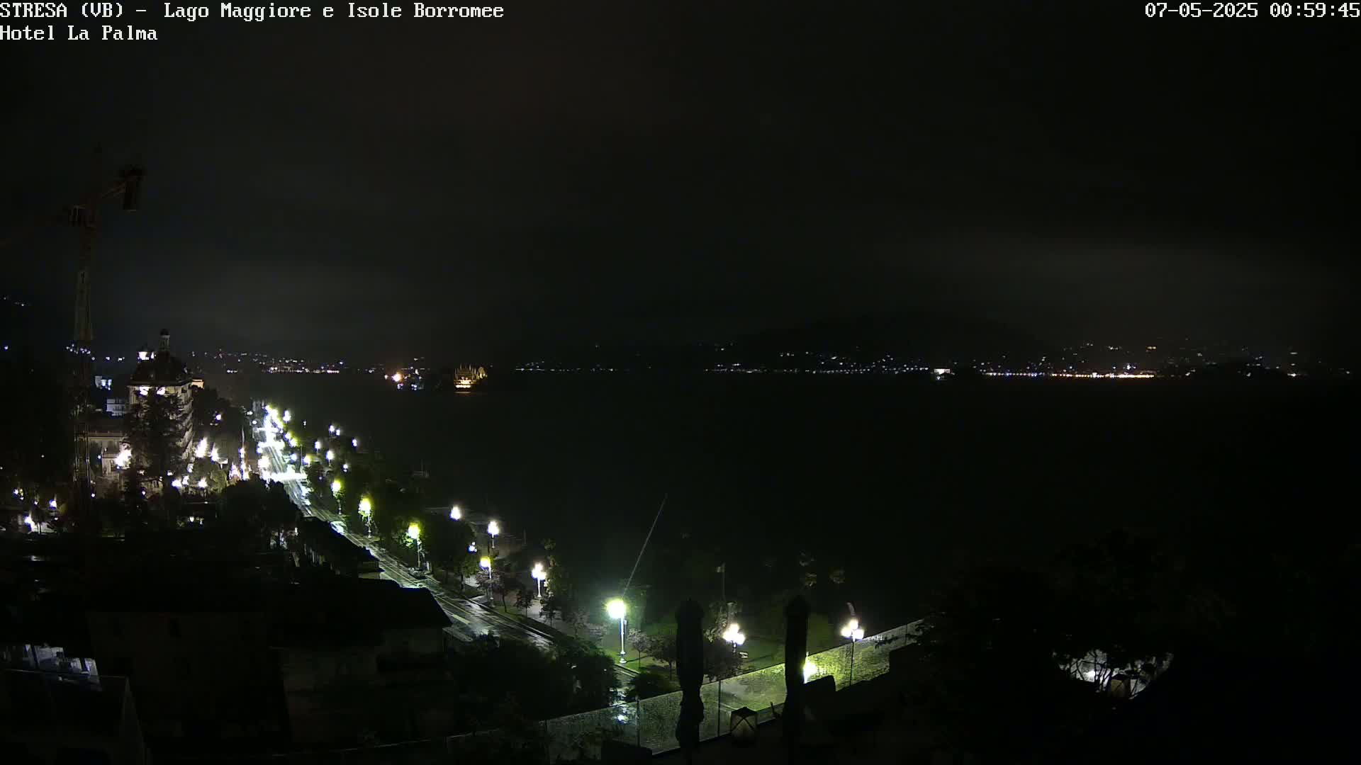 A nighttime, rainy view of a city and lake, with a road lined with lights leading towards a large body of water and distant city lights.
