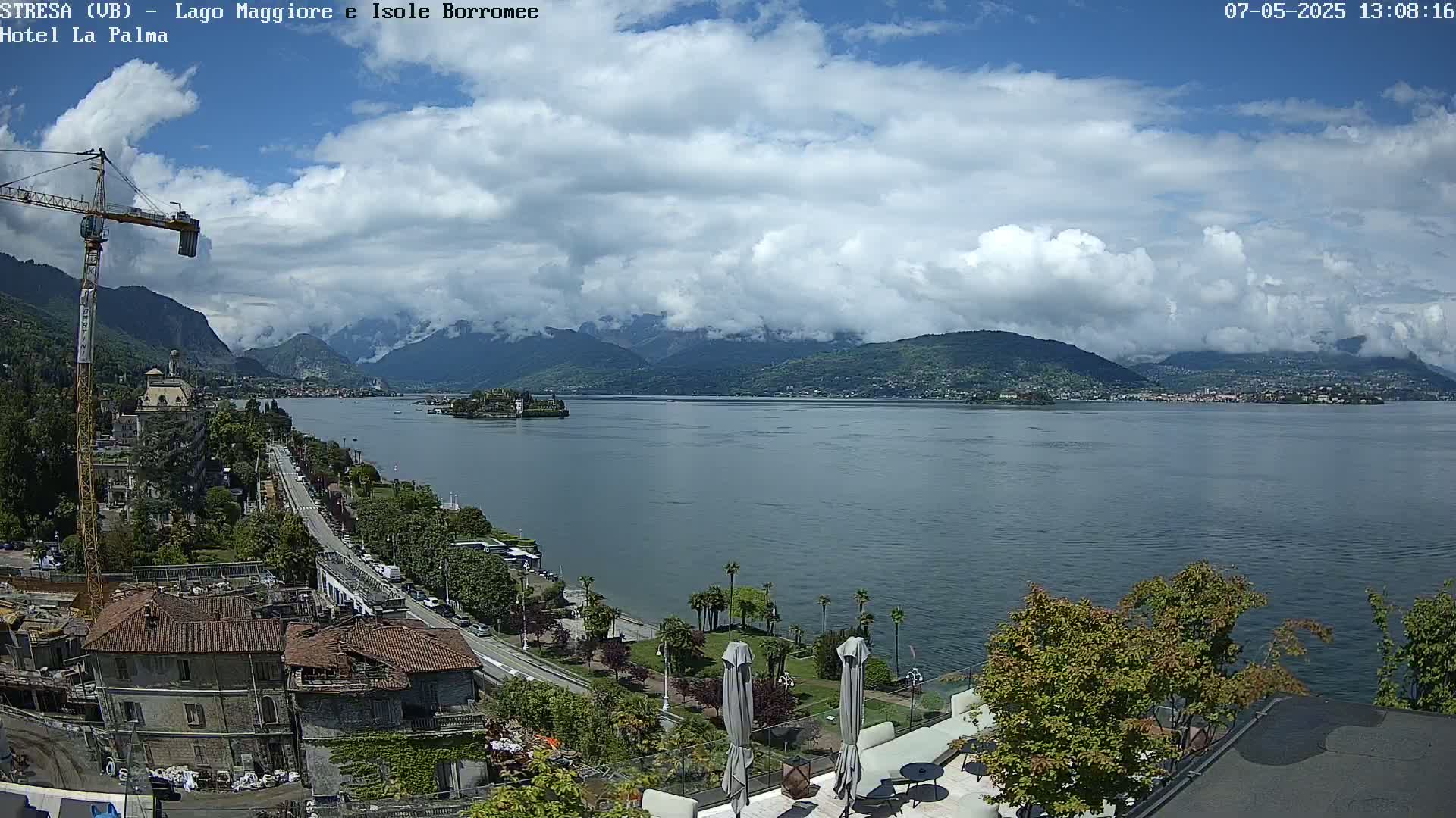 A partly cloudy day overlooking a large lake, with mountains in the background, a town along the lakeshore, and a construction crane visible near buildings under renovation.