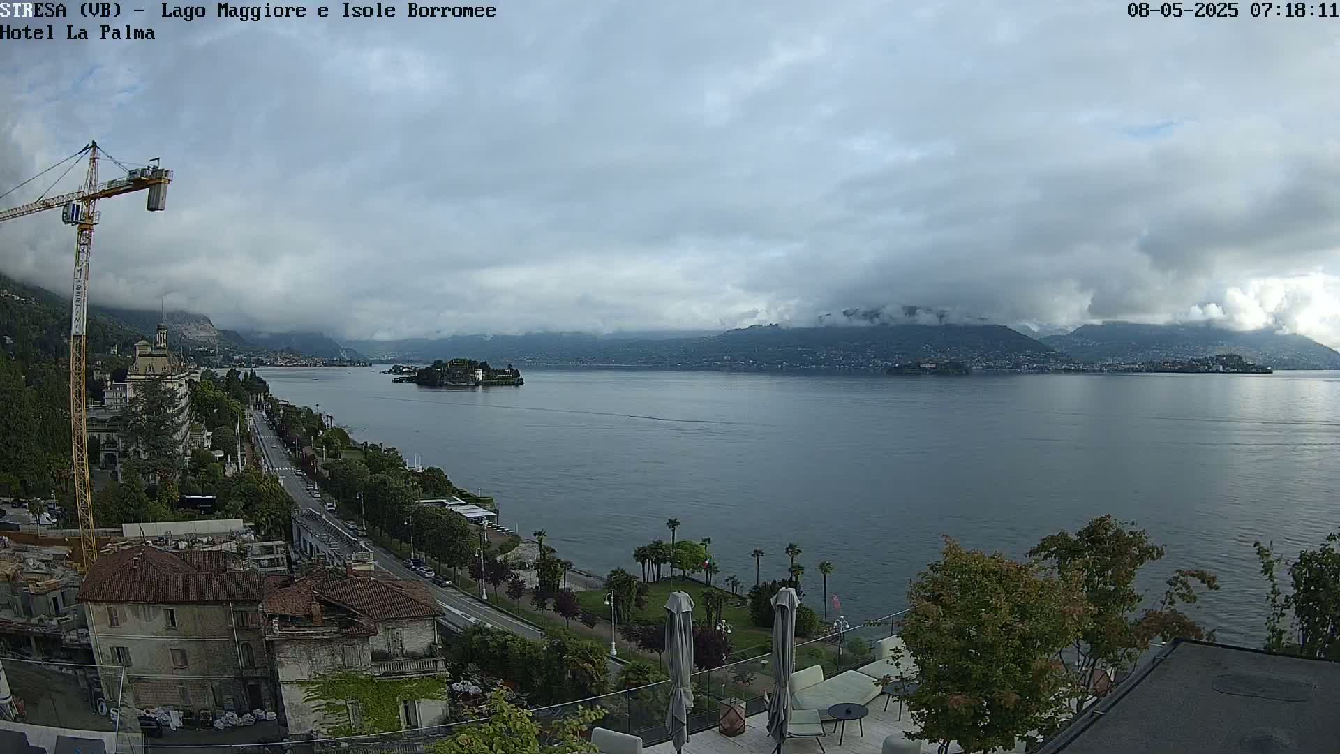 A partly cloudy day reveals a lake view with distant mountains, islands, a shoreline road lined with buildings, and a construction crane.