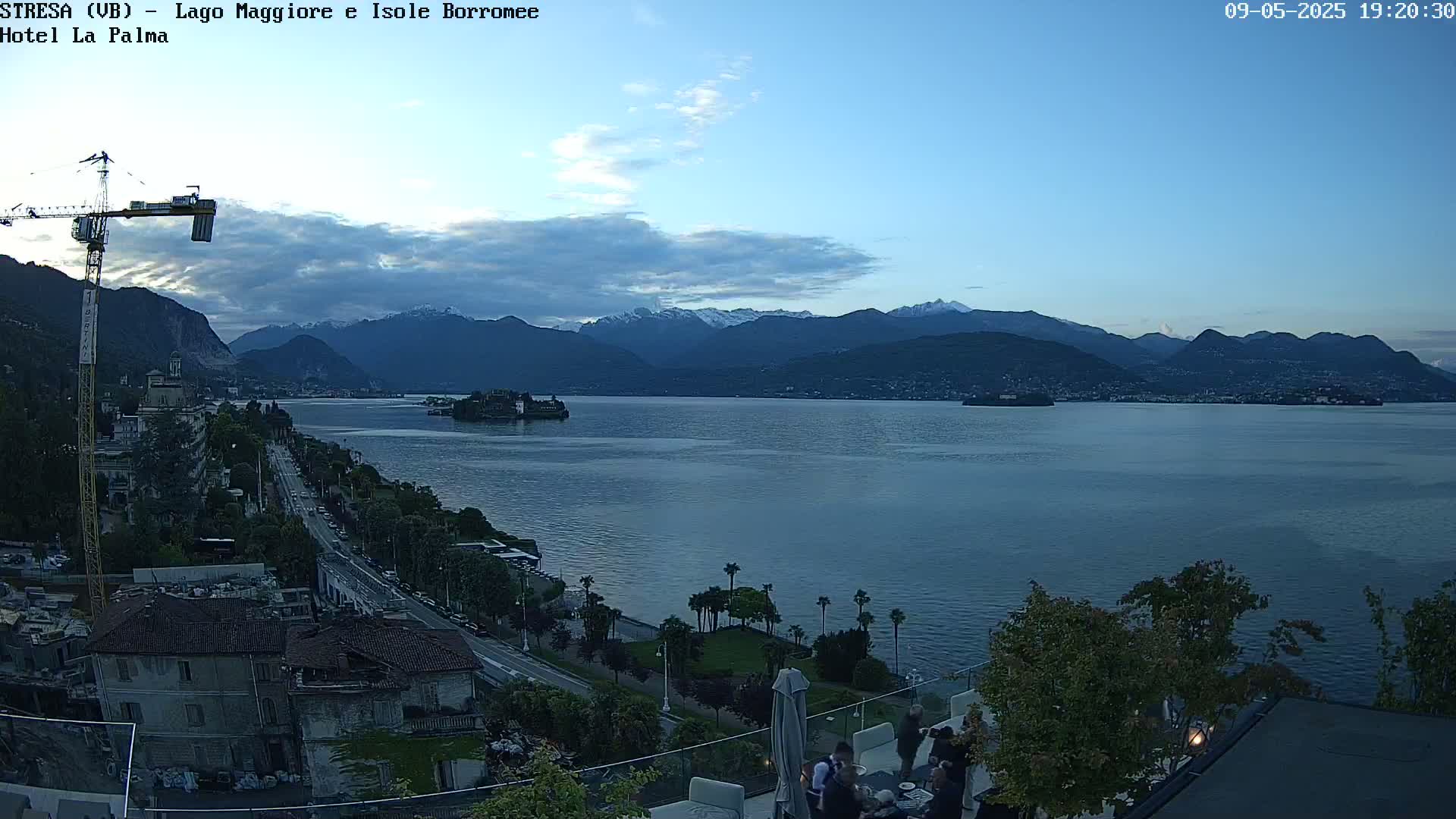 A twilight view of a lake with snow-capped mountains in the background, a town along the shoreline, and a crane visible near buildings.