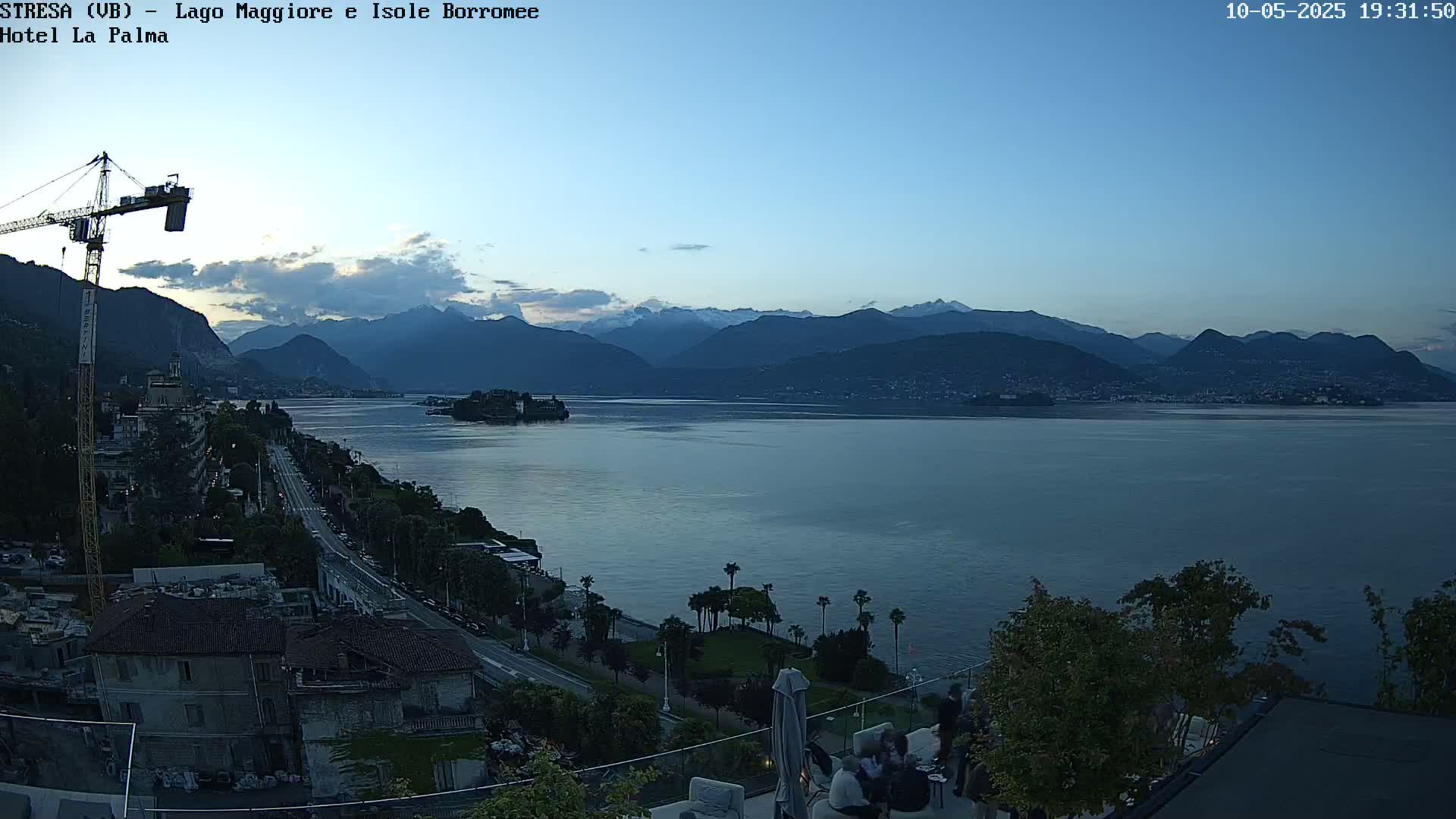 An elevated view of a calm lake at dusk, with mountains in the background, a town along the shoreline, and a few people sitting outside at a patio.