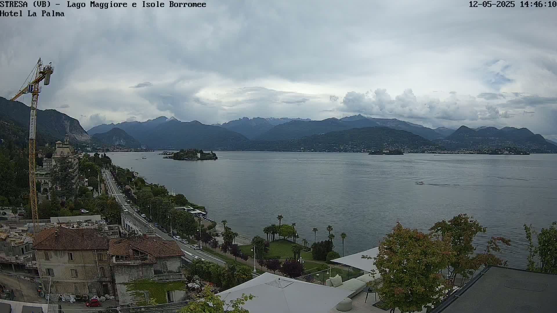 A wide, high-angle view shows a large lake with mountains in the background under a cloudy sky, a town and road along the lakeshore, and a crane in the foreground.