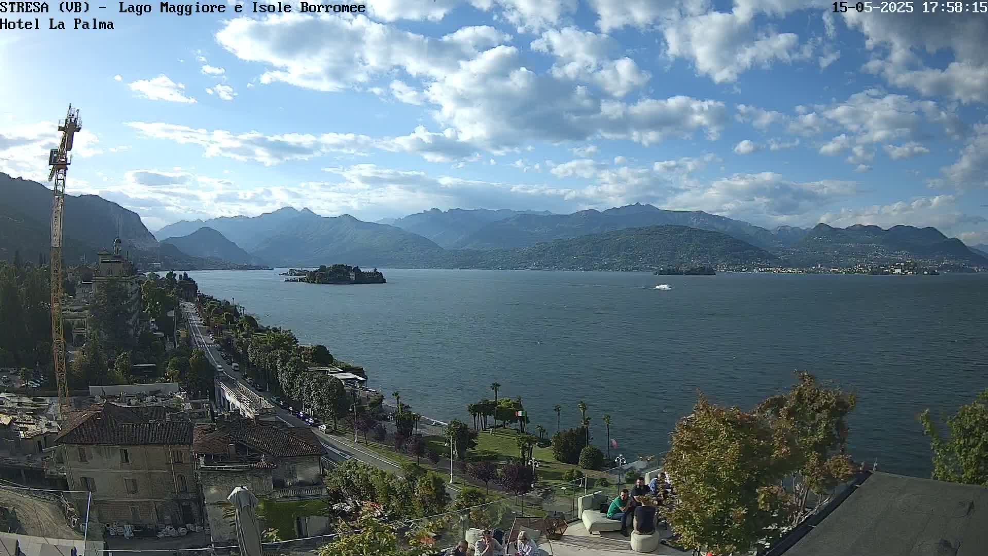 A partly cloudy day reveals a picturesque view of a lake with mountains in the background, a small island in the middle distance, and people gathered on a patio overlooking the water.