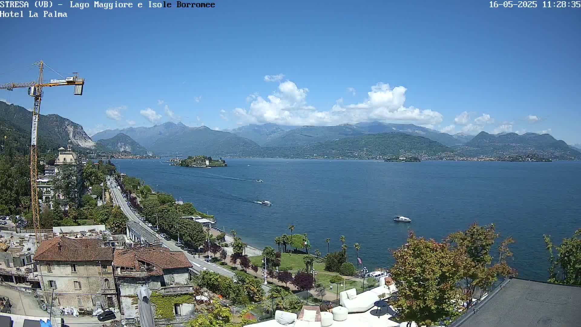 A partly cloudy sunny day overlooks a lake with several boats, a shoreline with buildings and trees, and mountains in the distance.