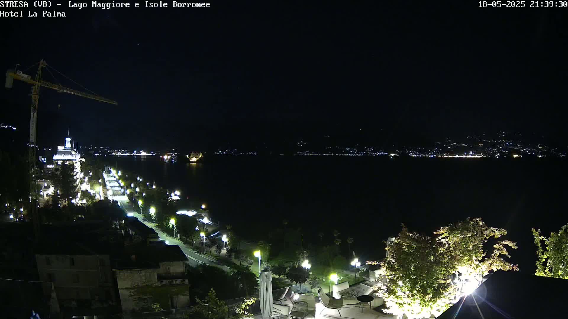 A nighttime, elevated view shows a city street lit by lamps extending toward a body of water with a distant, faintly lit shoreline under a clear, dark sky.
