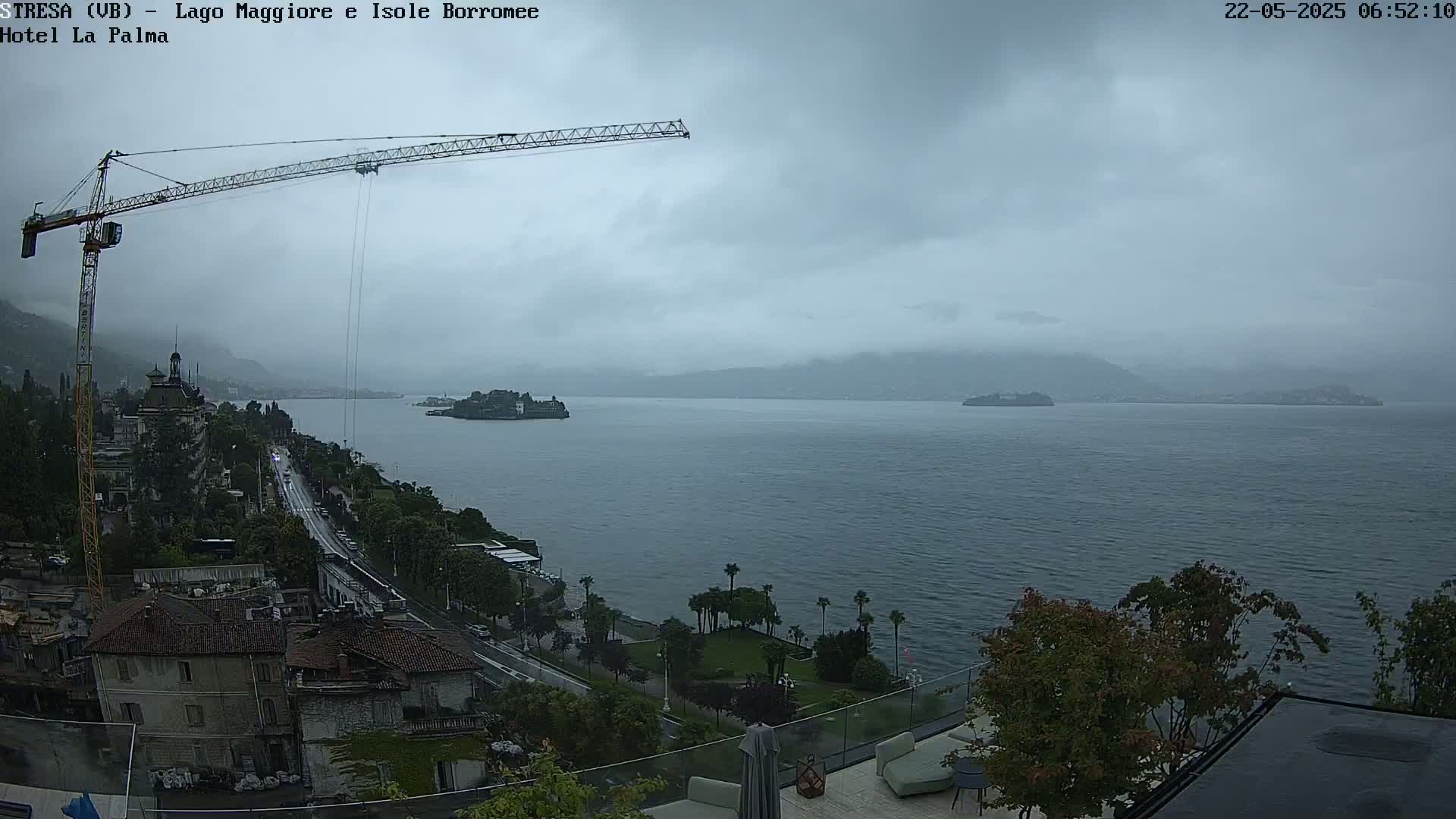 A large body of water is seen under an overcast sky, with a crane, buildings, and trees visible in the foreground.
