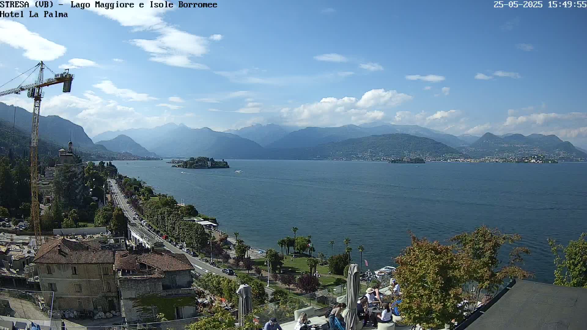 A partly cloudy sunny day overlooking a large lake with mountains in the distance, a town, and people seated at an outdoor patio.