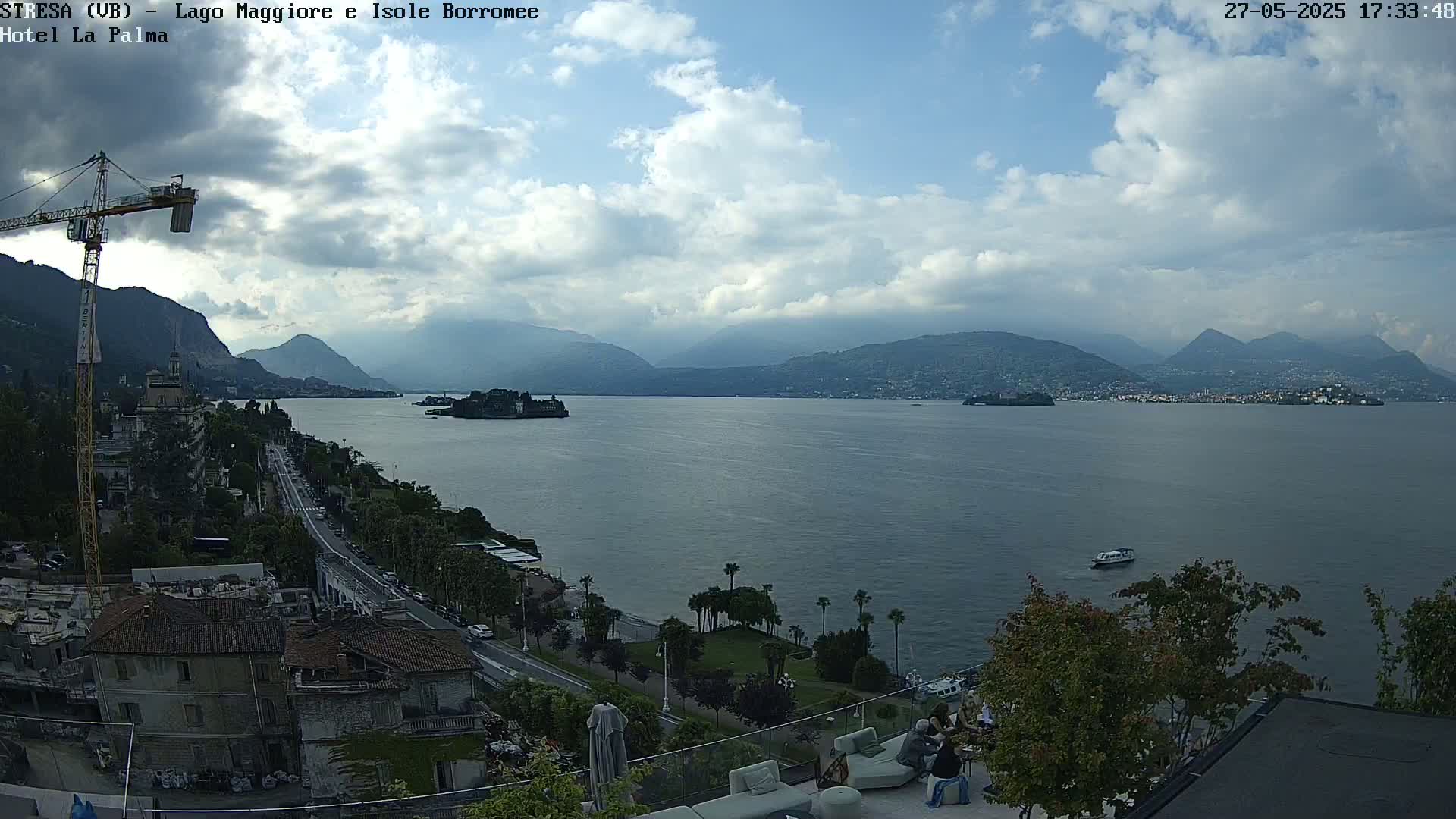 A partly cloudy day overlooking a large lake with mountains in the distance, a road and buildings along the shoreline, and several people sitting on an outdoor patio.