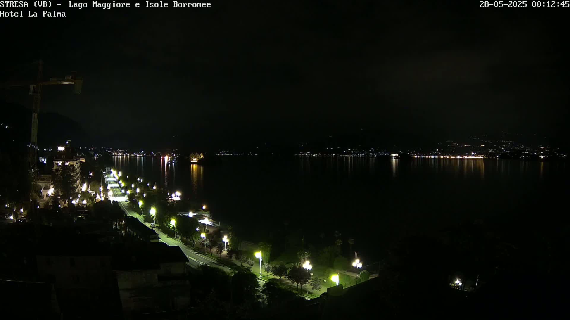 A nighttime, aerial view shows a dimly lit lakeside road with streetlights, leading to a dark body of water reflecting distant lights across its surface.