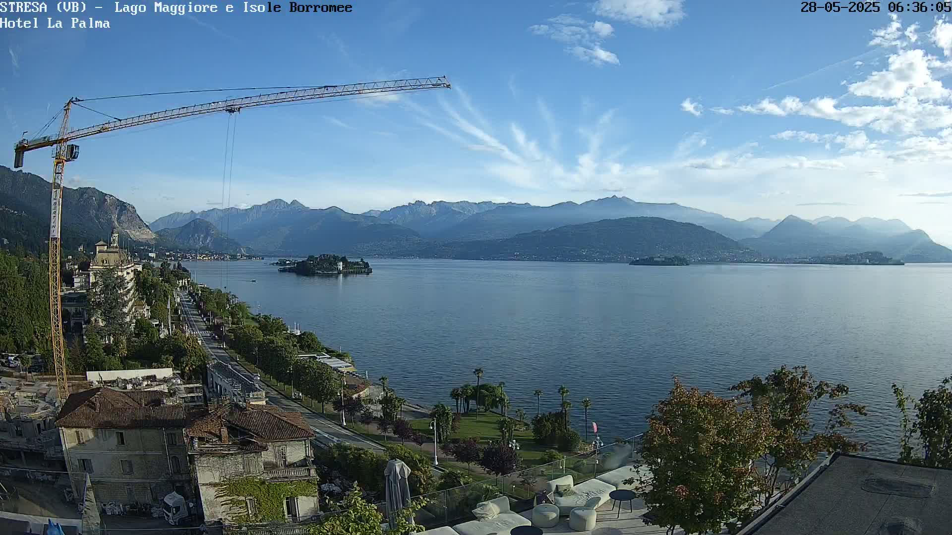 A large body of water stretches toward hazy mountains under a partly cloudy sky, with a crane, buildings, and vegetation visible in the foreground.