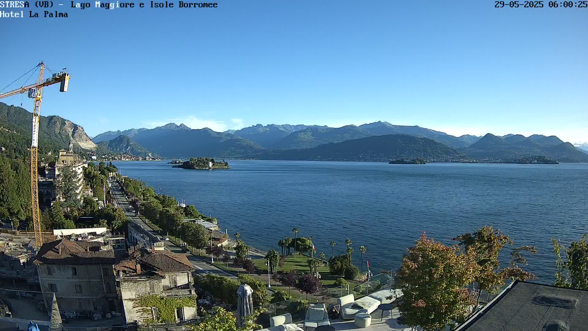 A clear, sunny day overlooking a calm lake with mountains in the background, a town visible along the lakeshore, and a construction crane near buildings.