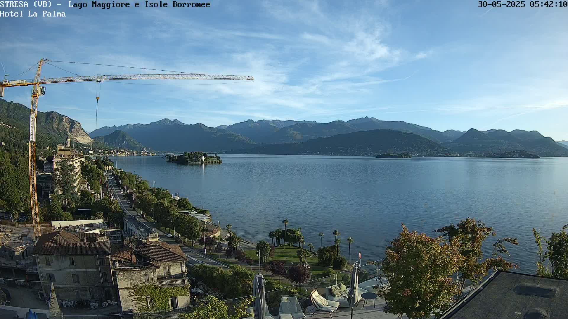A large lake with mountains in the background is seen from an elevated vantage point on a clear, sunny day; a crane is visible in the foreground near buildings and vegetation.
