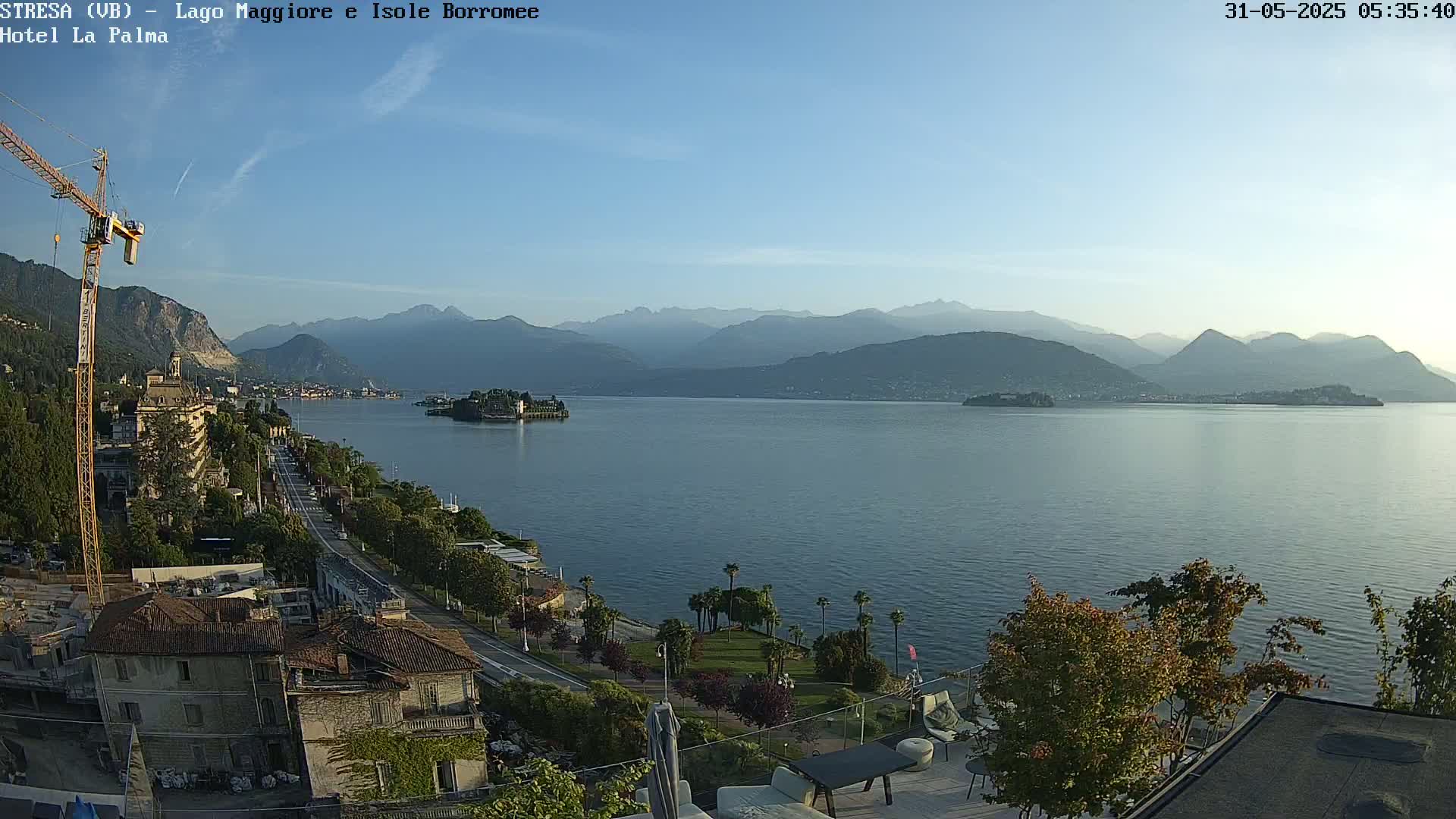 A tranquil lake scene under a clear sky, with mountains in the distance, a small island in the middle of the lake, and buildings and vegetation along the shoreline.