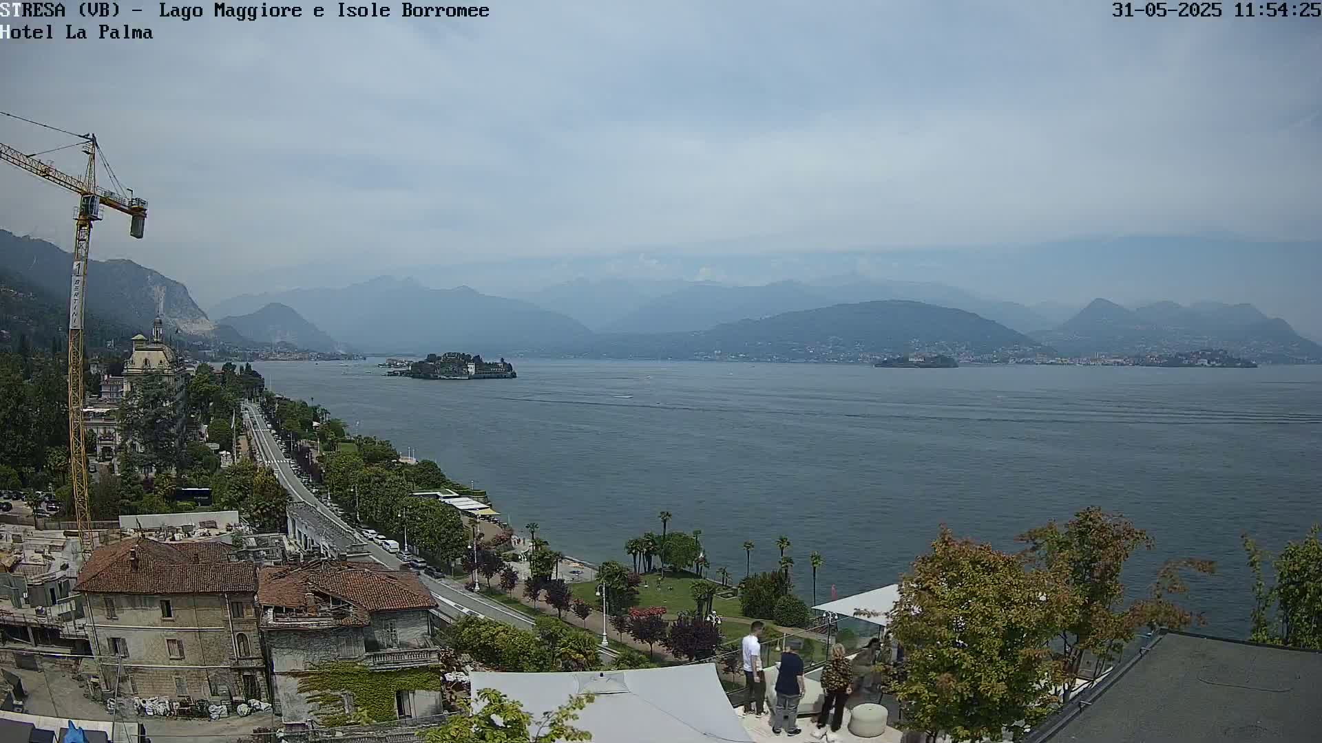 A partly cloudy day overlooks a large lake with mountains in the distance, a shoreline with buildings and trees, and a few people gathered near a structure.