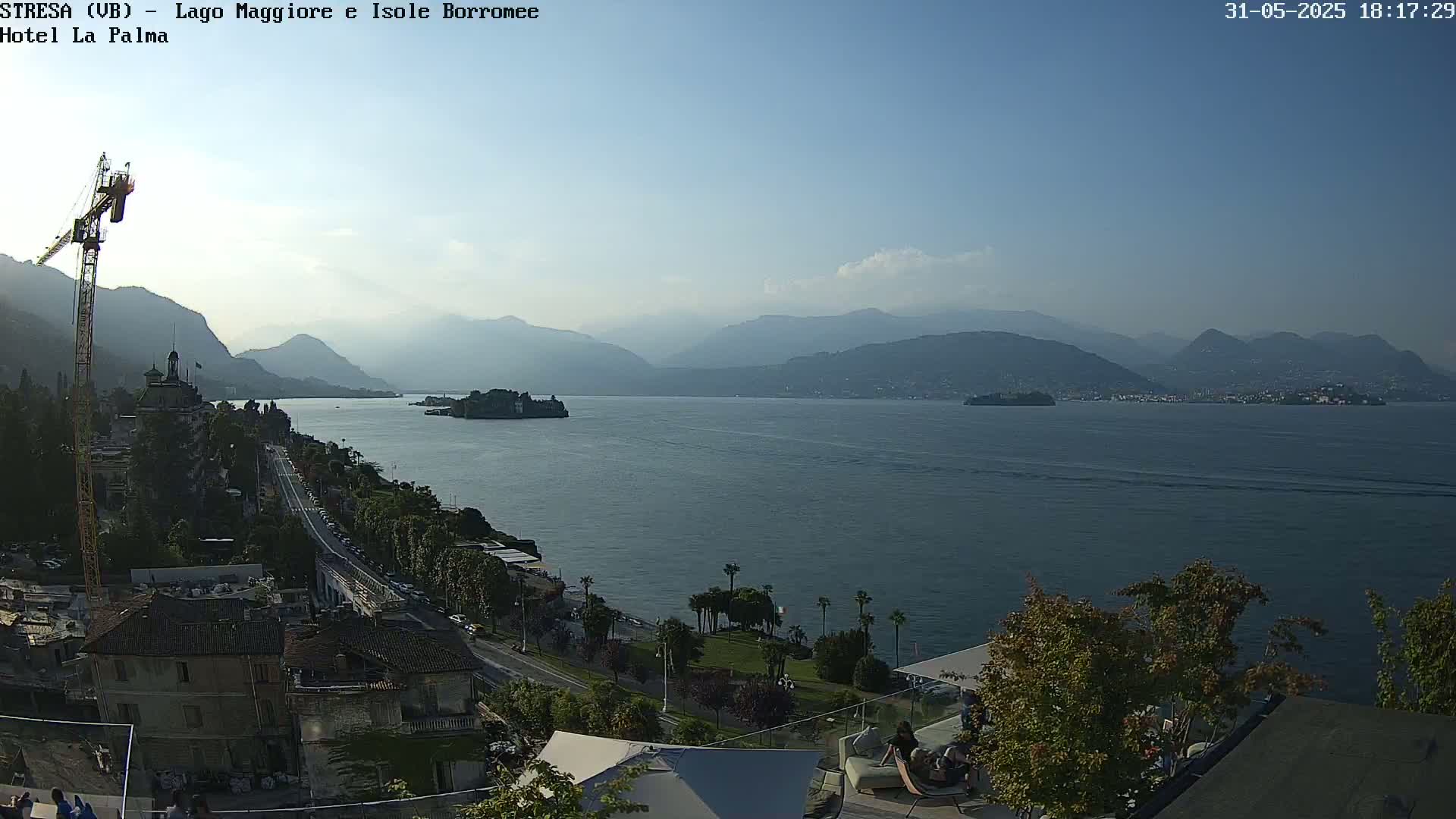 A clear, sunny day overlooking a large lake with islands and mountains in the distance, showing a town with a construction crane along the shoreline.