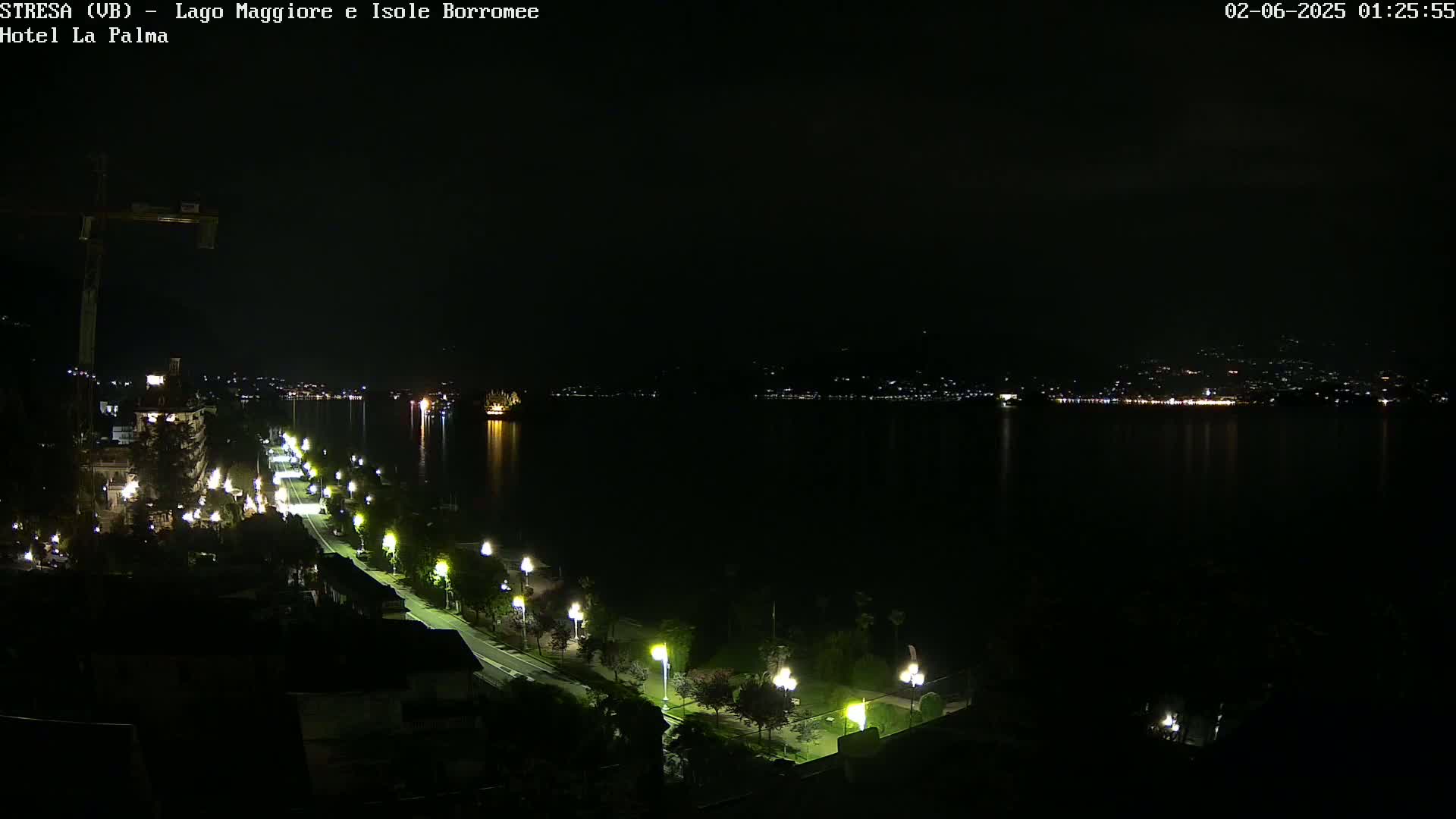 A nighttime view of a calm body of water with a dimly lit shoreline featuring a road lined with lamps, buildings, and a construction crane.